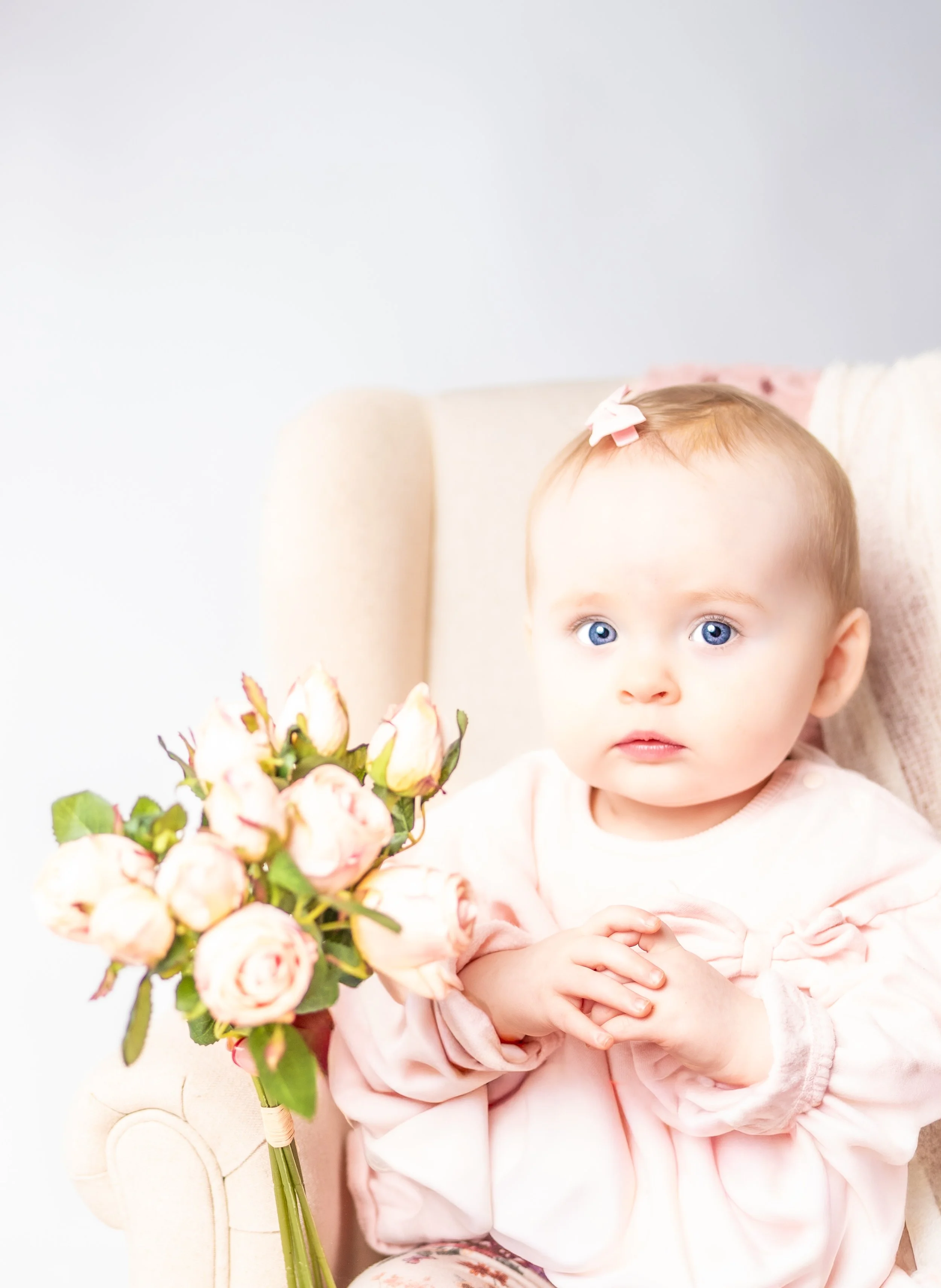 A young girl with blue eyes holding a bouquet of light pink roses, sitting on a beige armchair
