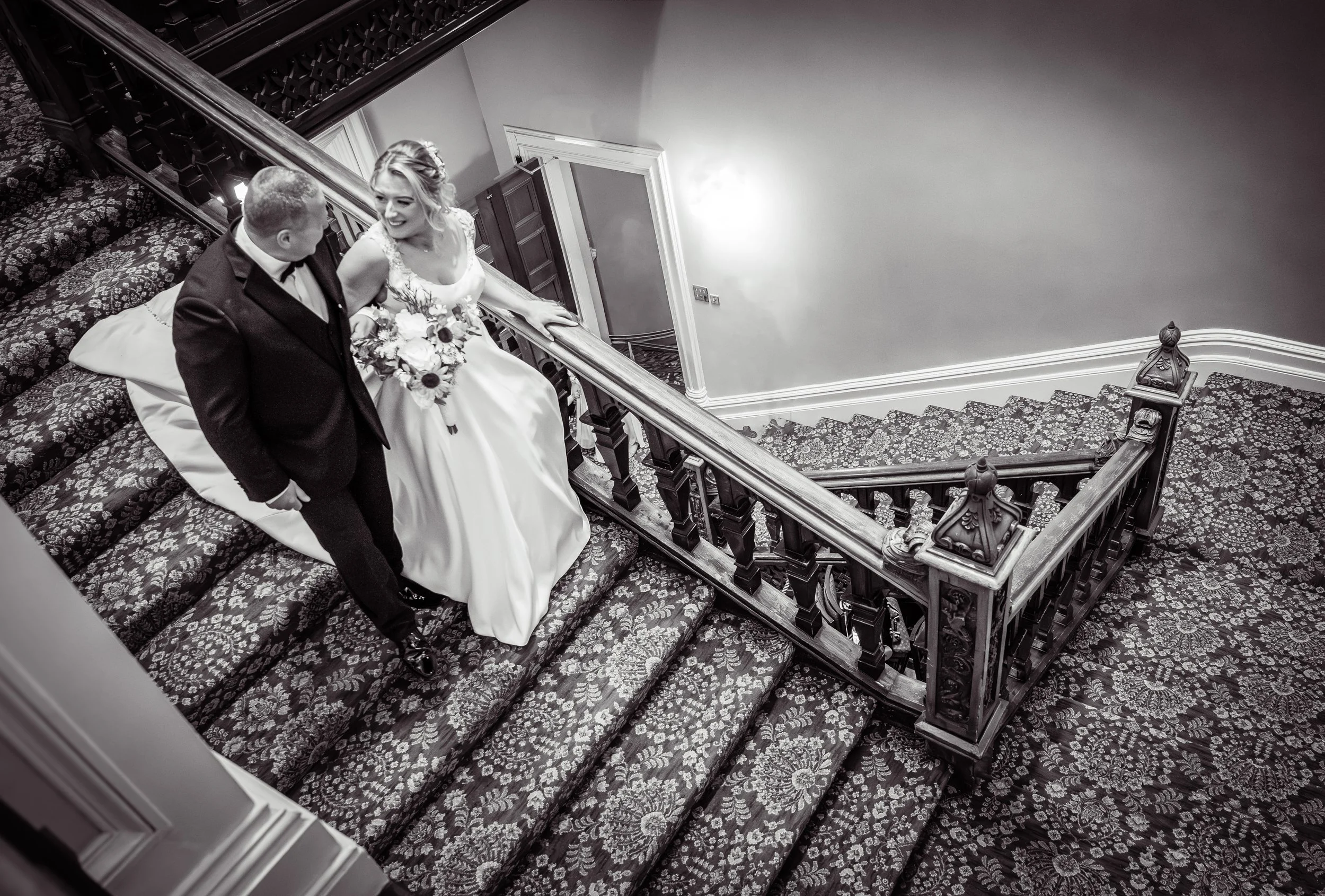 A black-and-white photo of a bride and groom walking up a staircase inside a building, with the bride holding a bouquet of flowers and smiling at the groom.