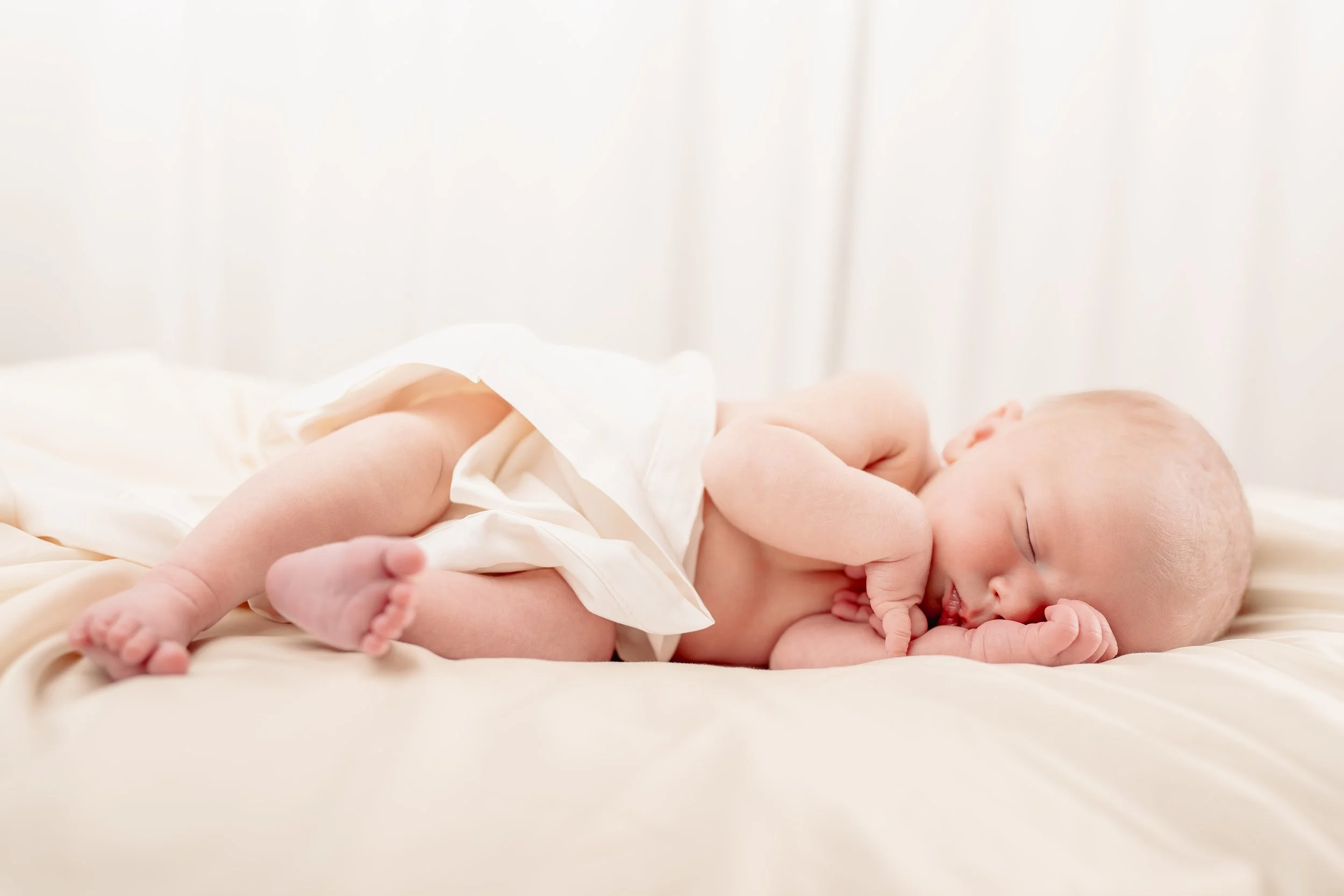 Close-up of a sleeping newborn baby lying on a soft surface, wearing a white cloth, with a gentle background.