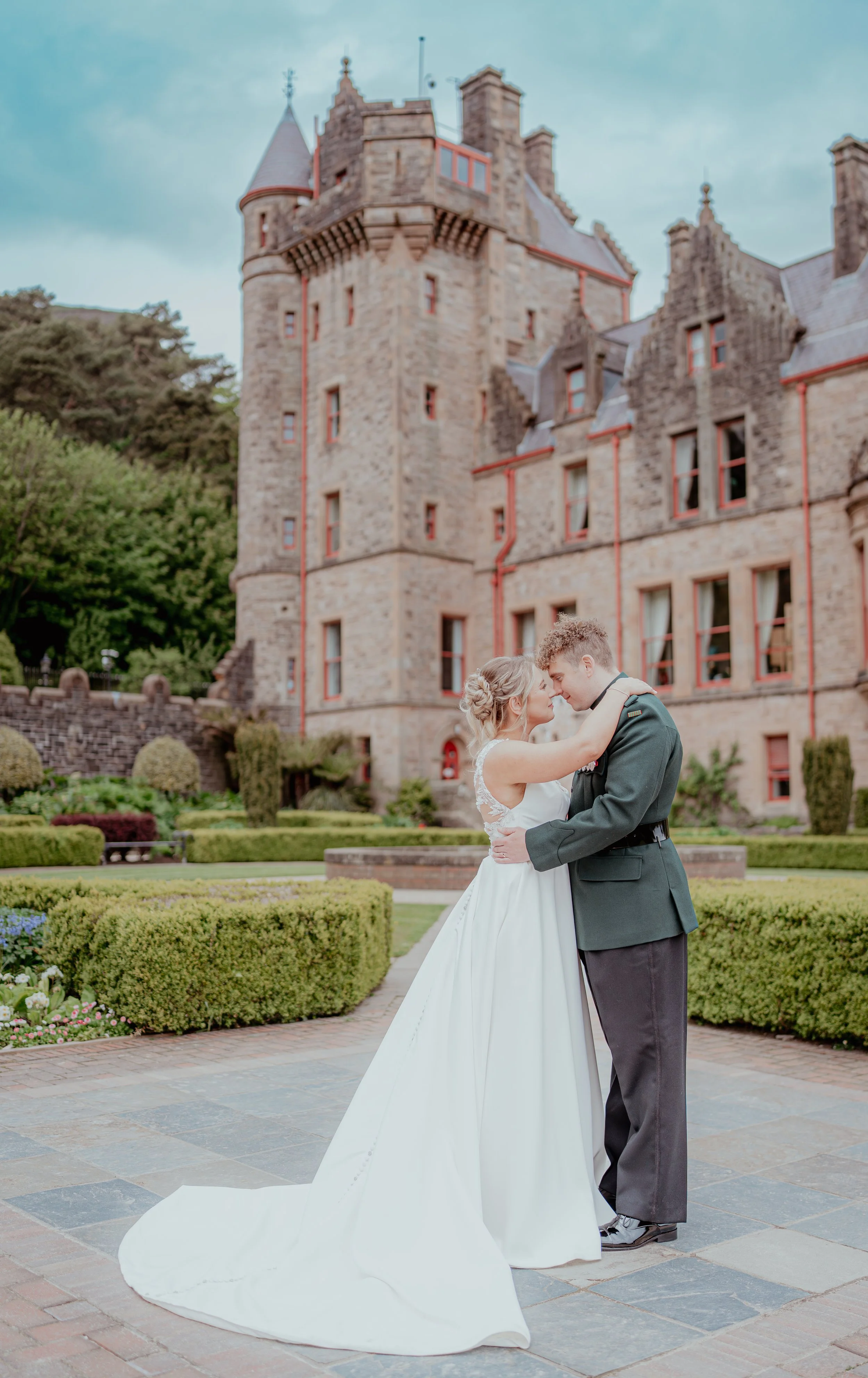 A newlywed couple in wedding attire, embracing and looking into each other's eyes, standing on a paved pathway in front of a historic stone castle with turrets and windows, surrounded by neatly trimmed garden hedges and lush greenery.