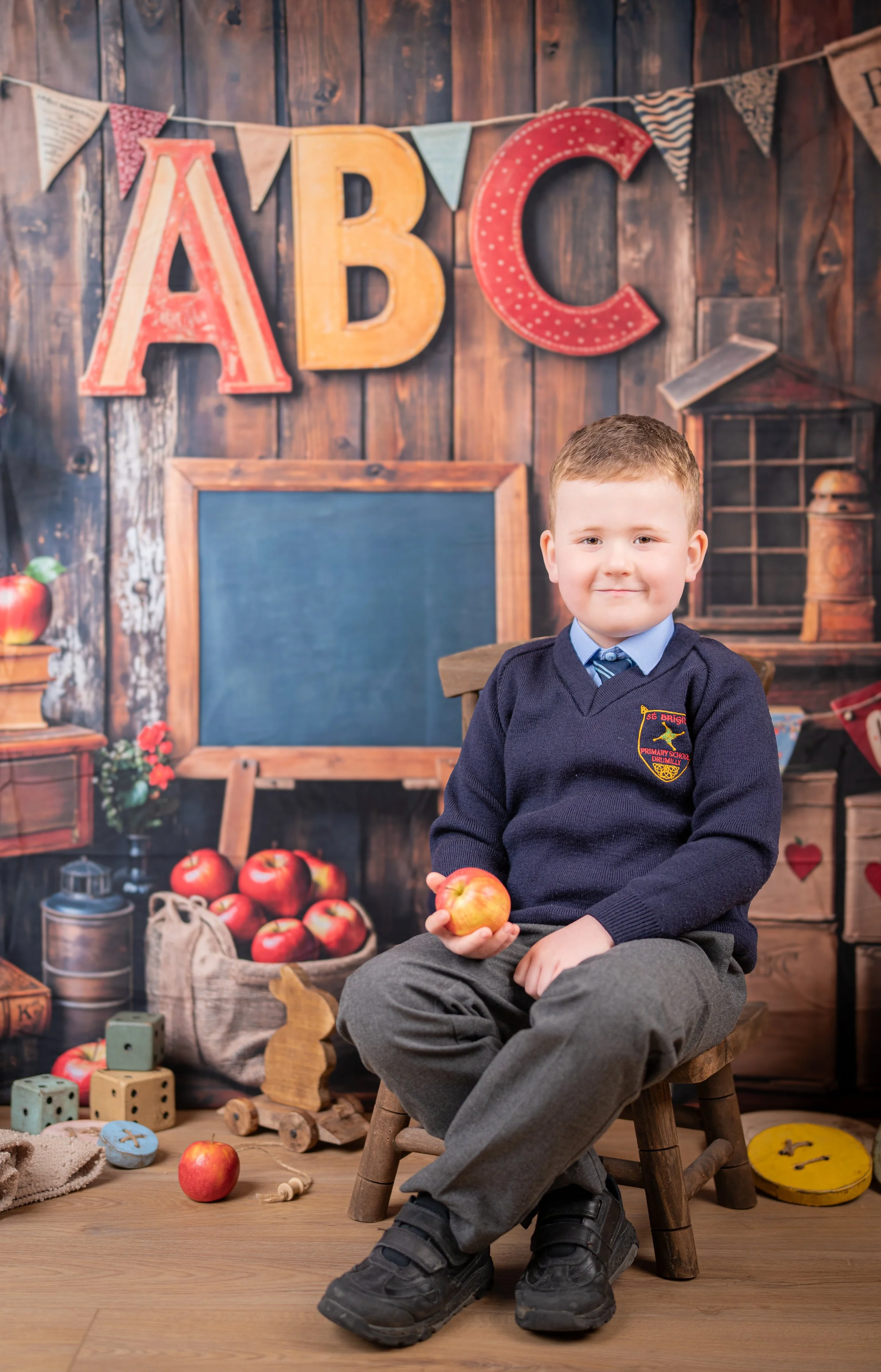A young boy in a school uniform sitting on a small wooden stool in front of a classroom-themed backdrop, holding an apple, with colorful alphabet letters and educational decorations around.