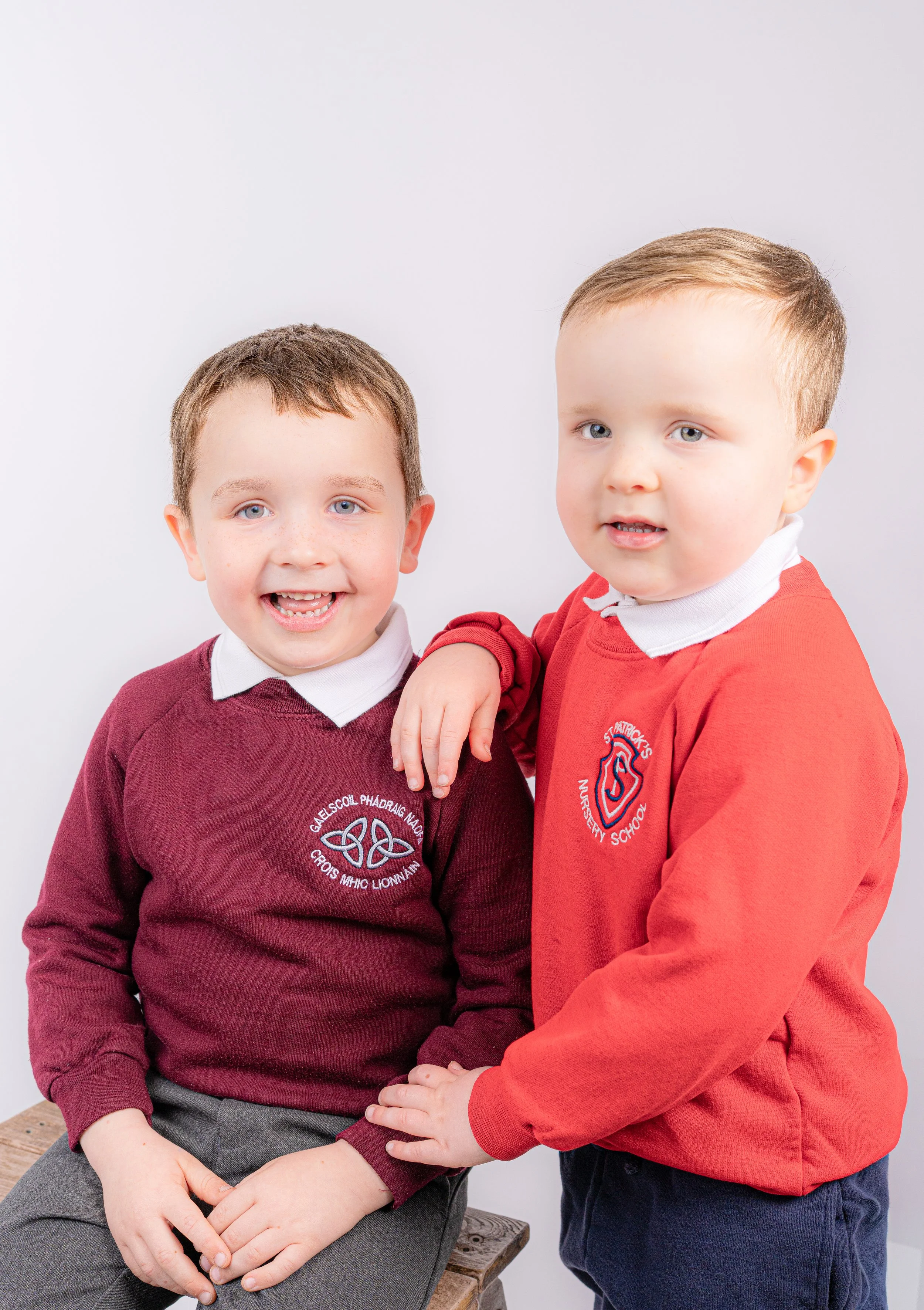 Two young boys in school uniforms, one sitting on a wooden stool and the other standing beside him with his arm on the sitting boy's shoulder, smiling at the camera against a plain white background.