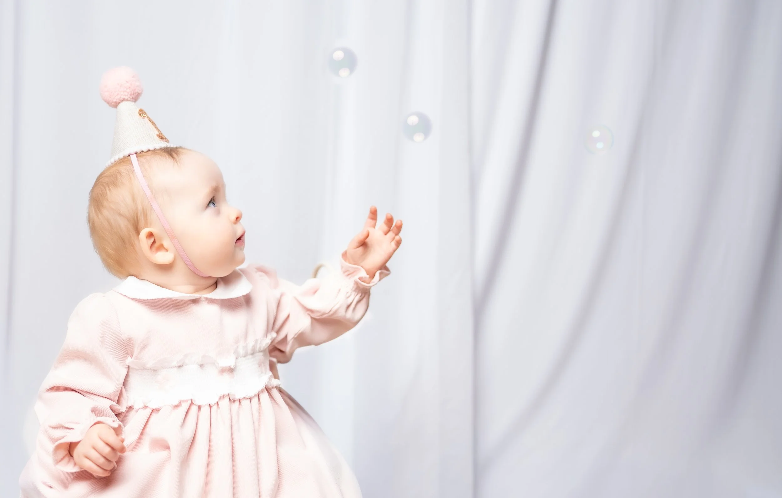 A young girl wearing a pink dress and a birthday hat, reaching out and looking at floating bubbles against a white curtain background.