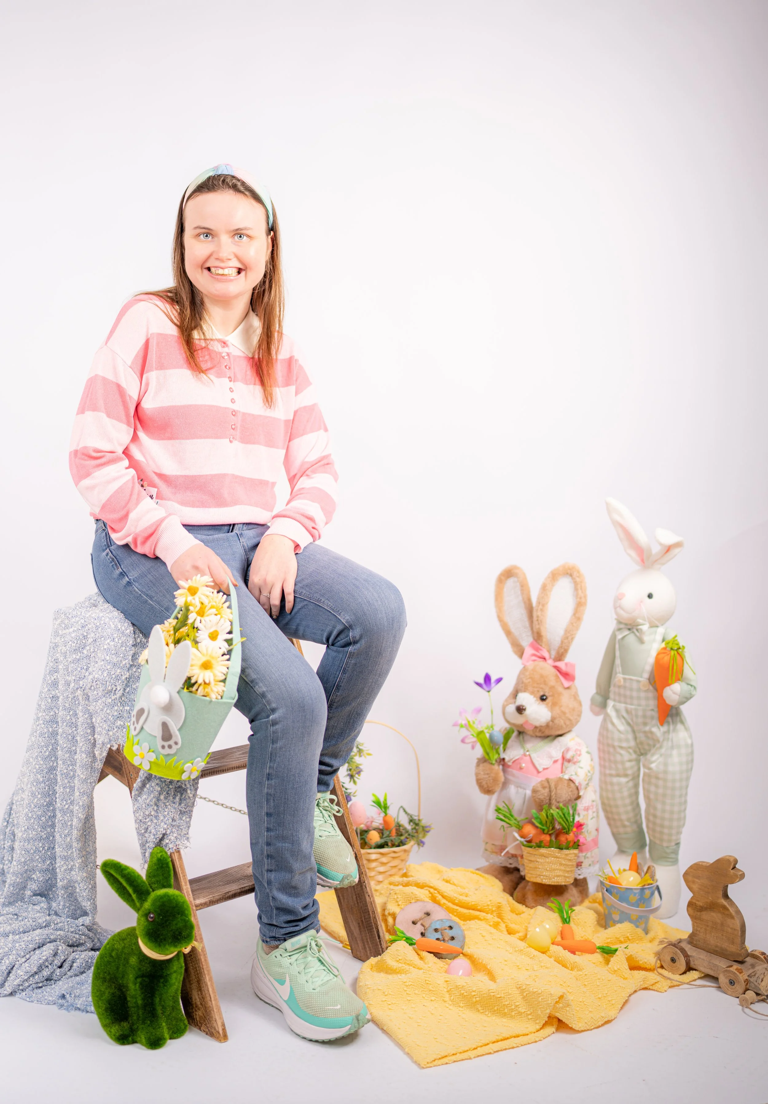 Woman sitting on a stool with Easter decorations, including stuffed bunnies, carrots, eggs, a green plush bunny, and baskets of flowers, in a bright studio with white background.