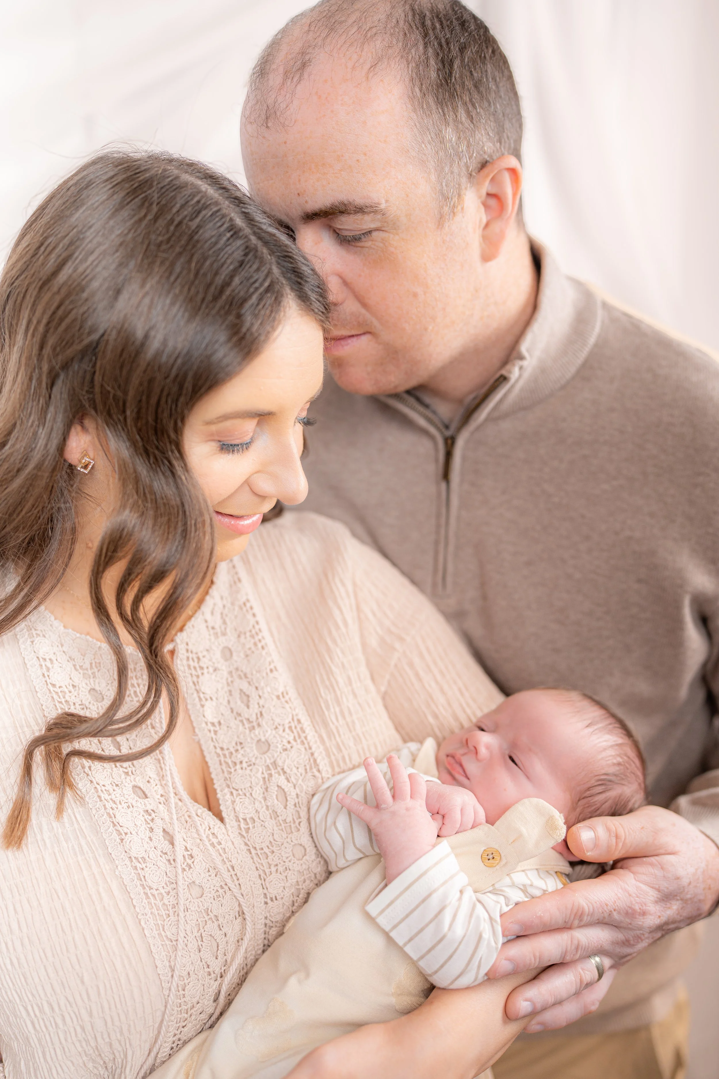 A family of three softly holding and gently looking at a sleeping baby, with the mother and father leaning close in a warm, intimate moment.