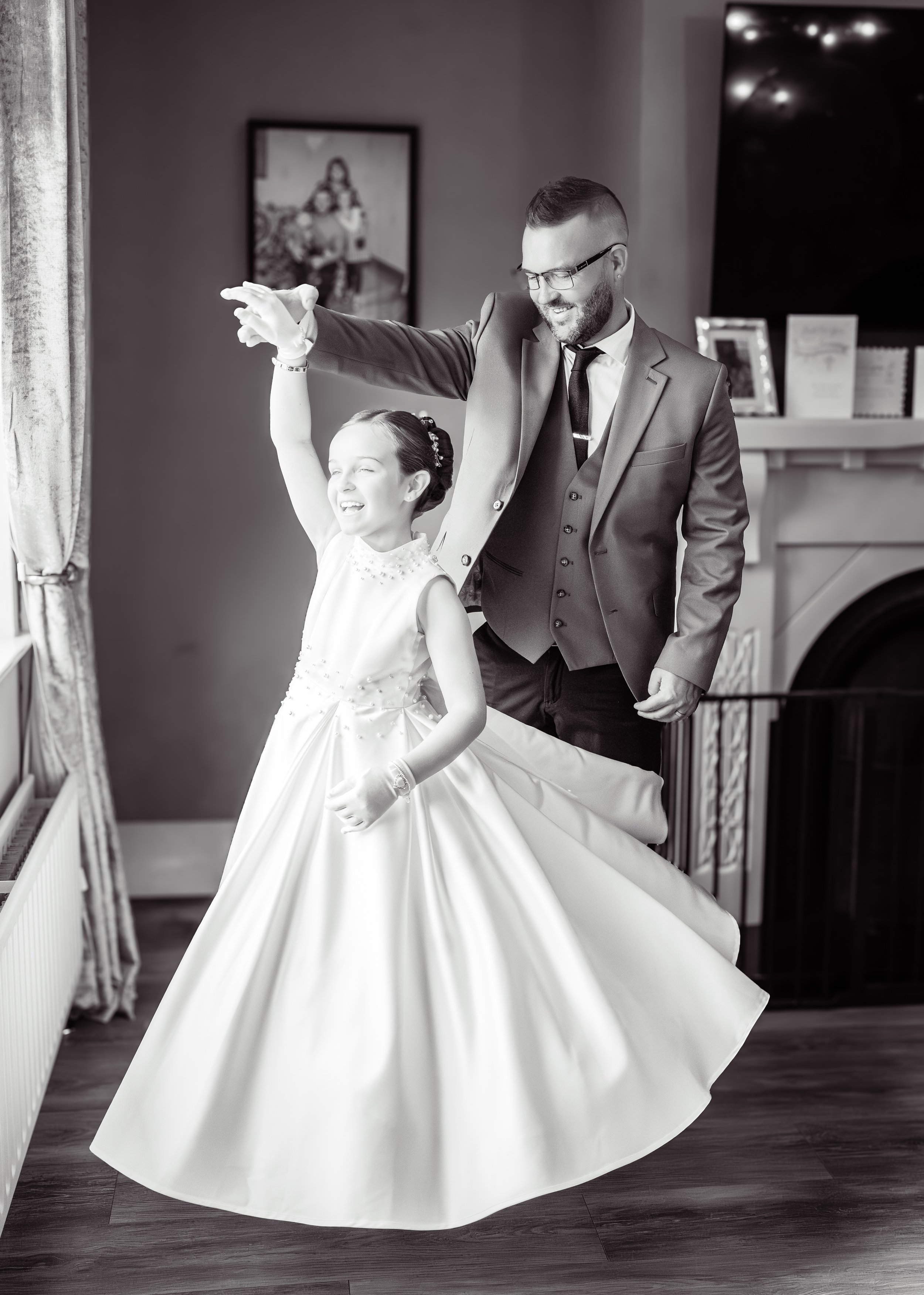 A man in a suit and glasses dancing with a young girl in a white dress, both smiling and enjoying the moment indoors.