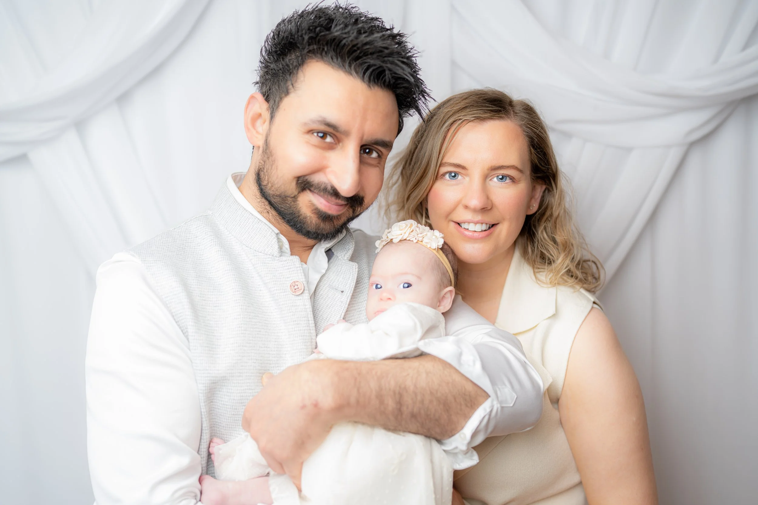 A happy family of three, including a father, mother, and their baby, smiling and posing together against a white backdrop.