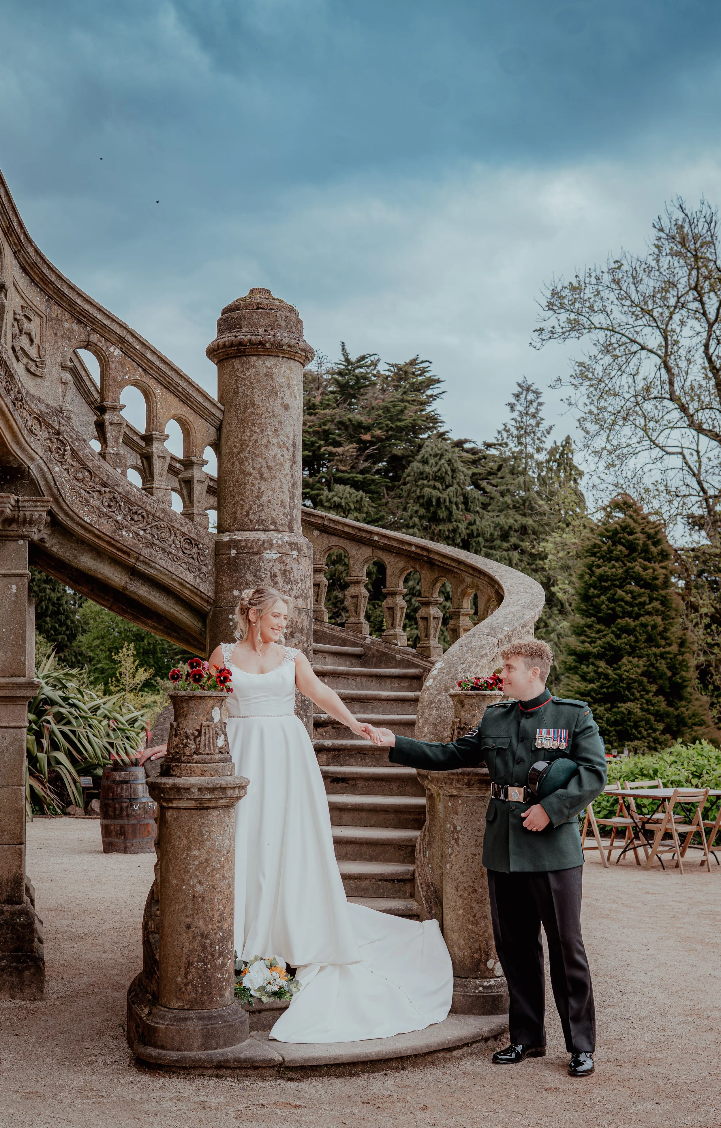 A bride in a white wedding dress holding hands with a groom dressed in a military uniform, standing on an outdoor stone staircase with columns and decorative railings, surrounded by greenery and trees, with cloudy sky overhead.
