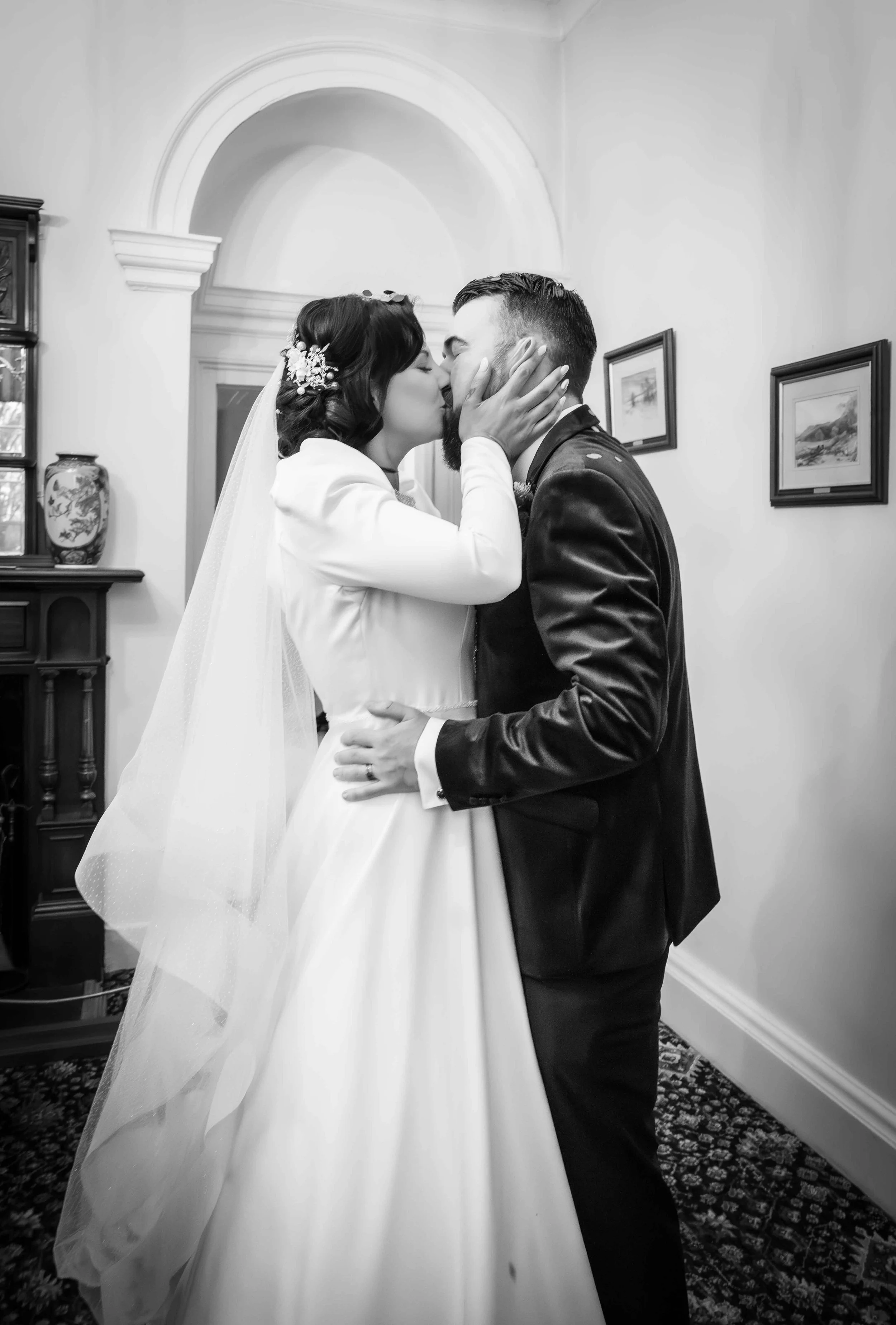 A bride and groom share a kiss indoors, with the bride holding the groom's face and the groom's hand around the bride's waist, in a black-and-white photo.