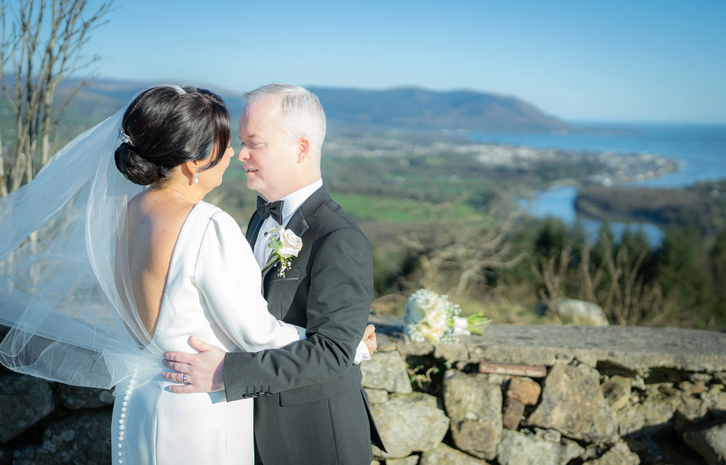 A bride and groom embrace outdoors on their wedding day, with scenic green hills, a river, and the ocean in the background.