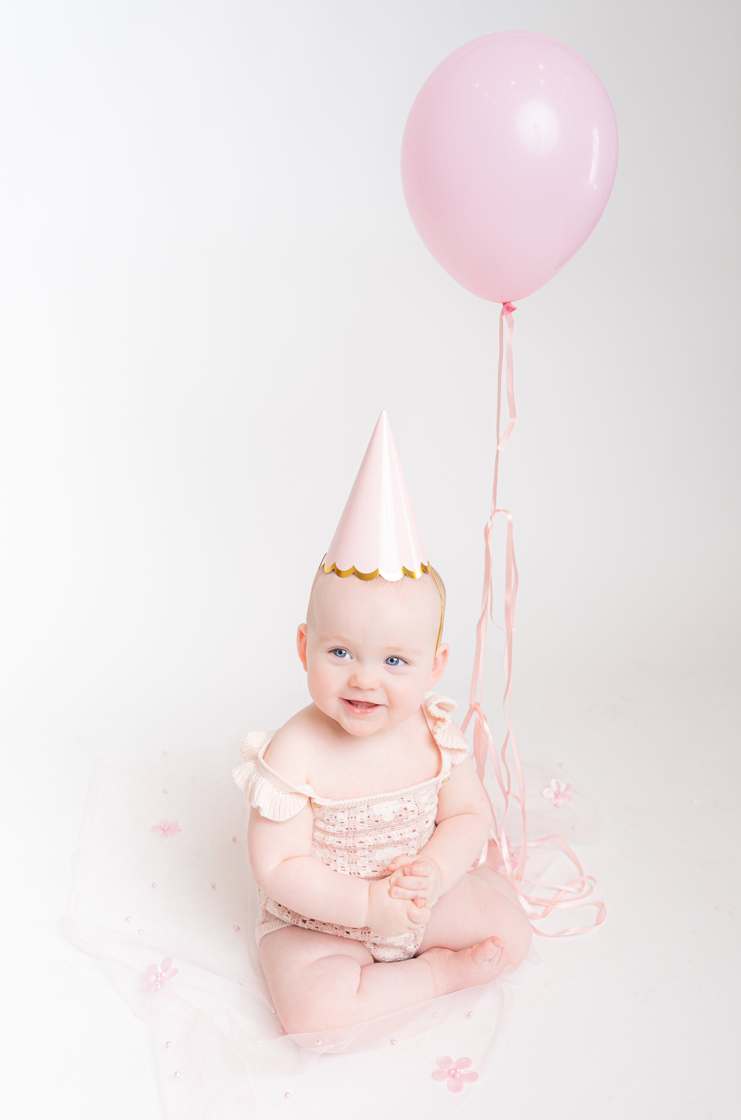 A smiling baby girl sitting on a white blanket, wearing a pink dress with ruffled sleeves. She has a pink party hat with gold trim on her head. Behind her, there is a pink helium balloon tied with pink ribbons. The blanket is decorated with pink flower-shaped embellishments, and the background is plain and white.