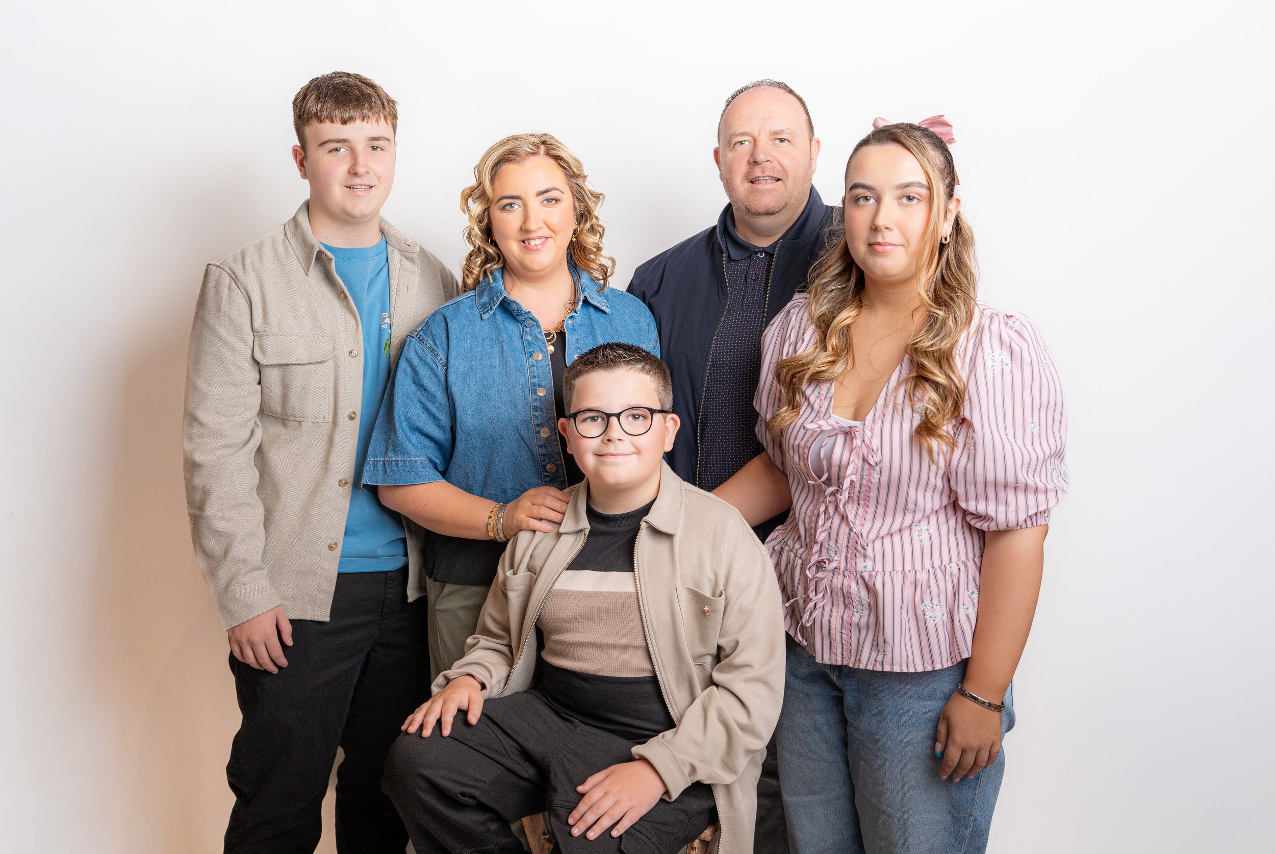 A family portrait with five members standing and sitting in front of a plain white background. They include two adults and three children, all smiling or looking at the camera.