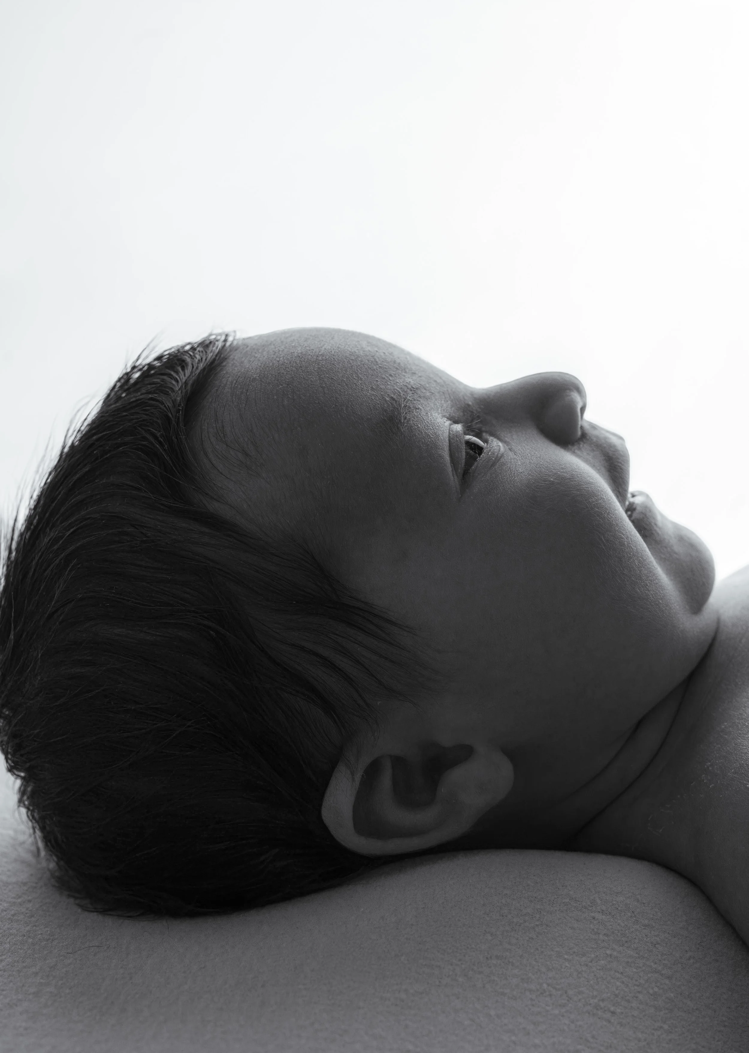 Close-up black and white photo of a woman's face lying on a surface, viewed from the side with eyes open and hair parted to one side.