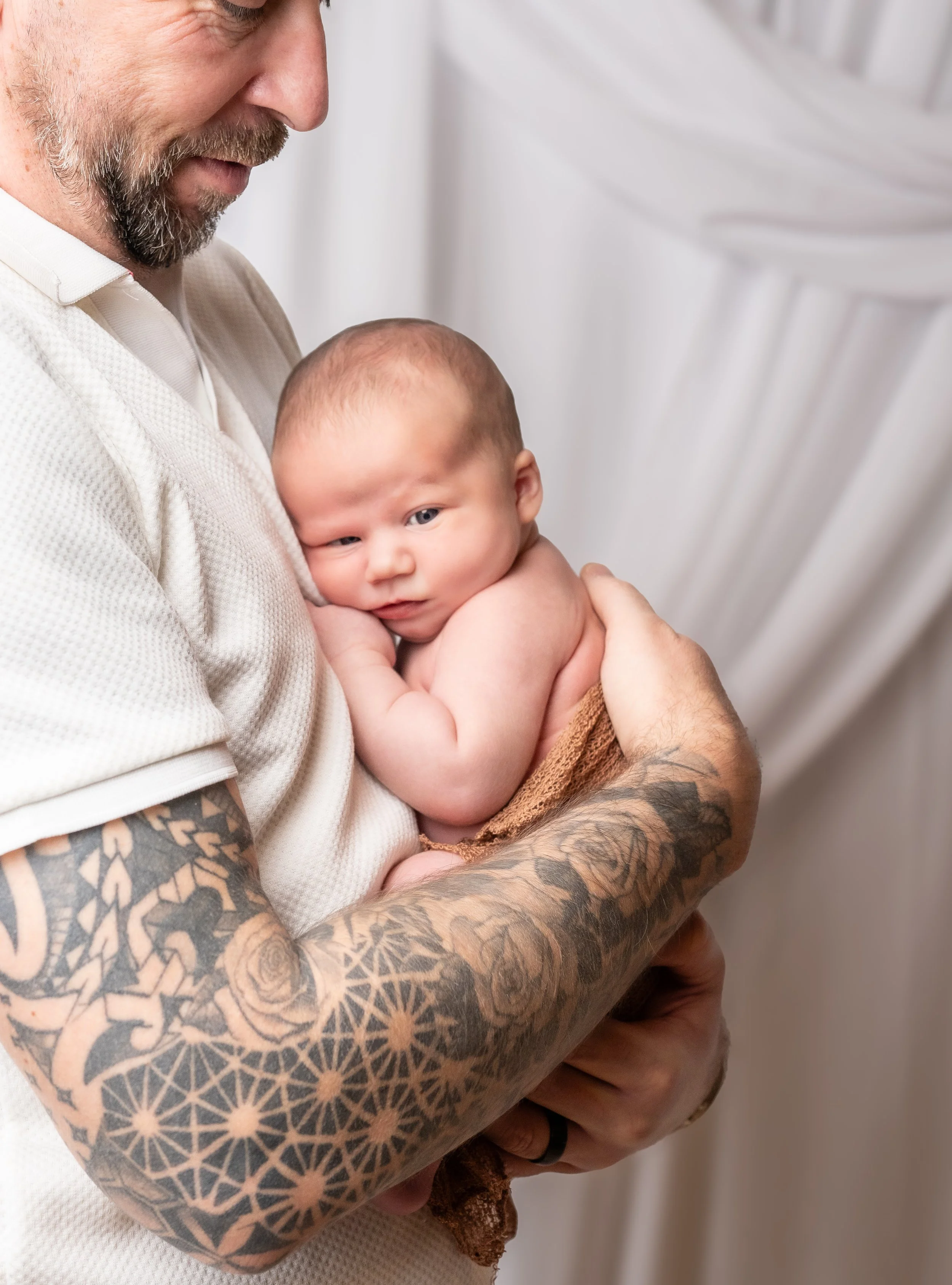 A man with tattoos on his arms holding a baby with a light complexion and a bald head, in front of a white background.