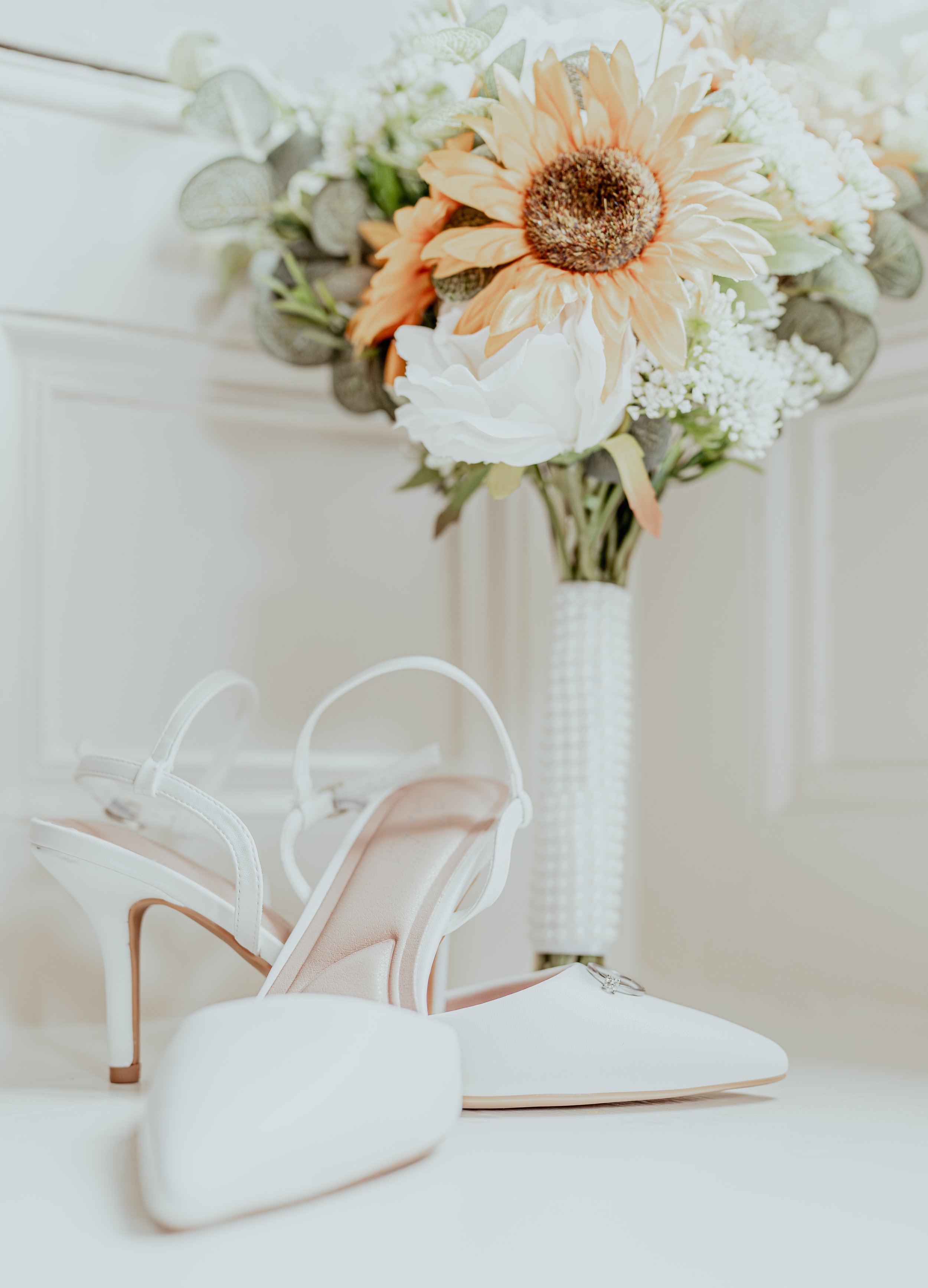 Close-up of a bridal bouquet with large peach and white flowers, greenery, and decorative elements, placed in a tall white textured vase. In the foreground, a pair of white and blush pink high-heeled shoes with ankle straps are visible on a white surface.