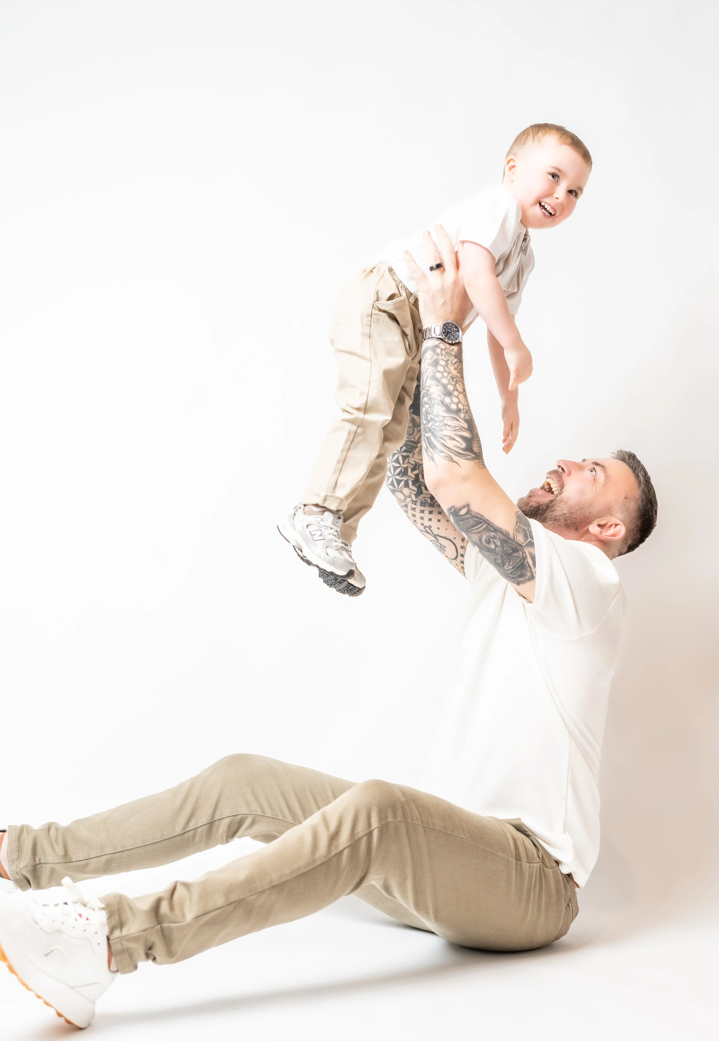 A man with tattoos sitting on the floor playing with a young boy, lifting him up in the air, both smiling joyfully.