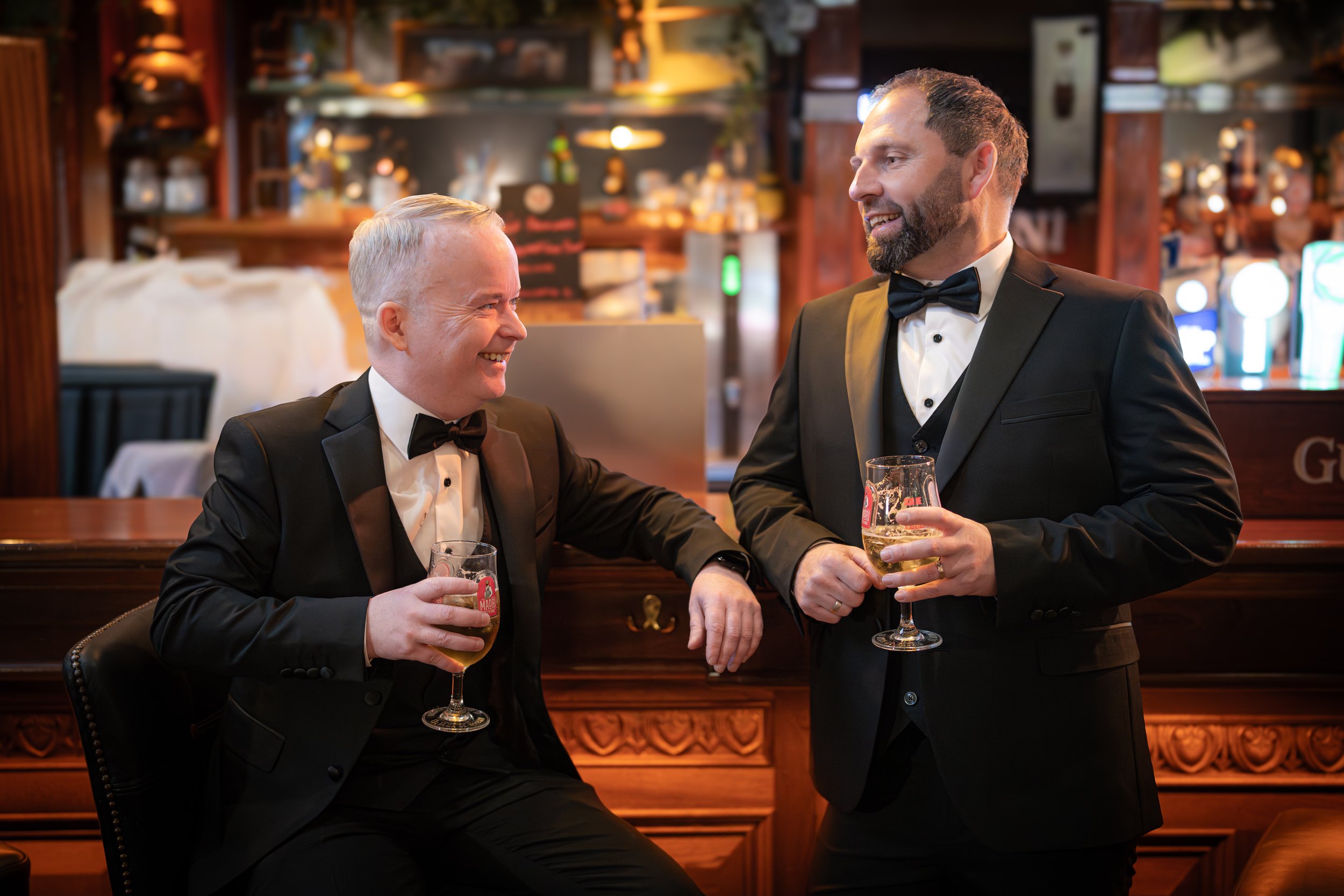 Two men in tuxedos holding glasses of beer, smiling and engaging in conversation inside a warmly lit bar or pub.