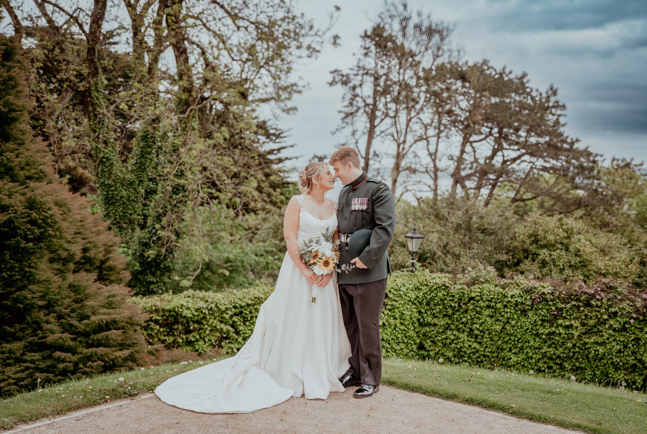 A bride and groom are standing face to face outdoors in a garden with trees and shrubs, gazing into each other's eyes, with the bride holding a bouquet of sunflowers and greenery and the groom in a military uniform.