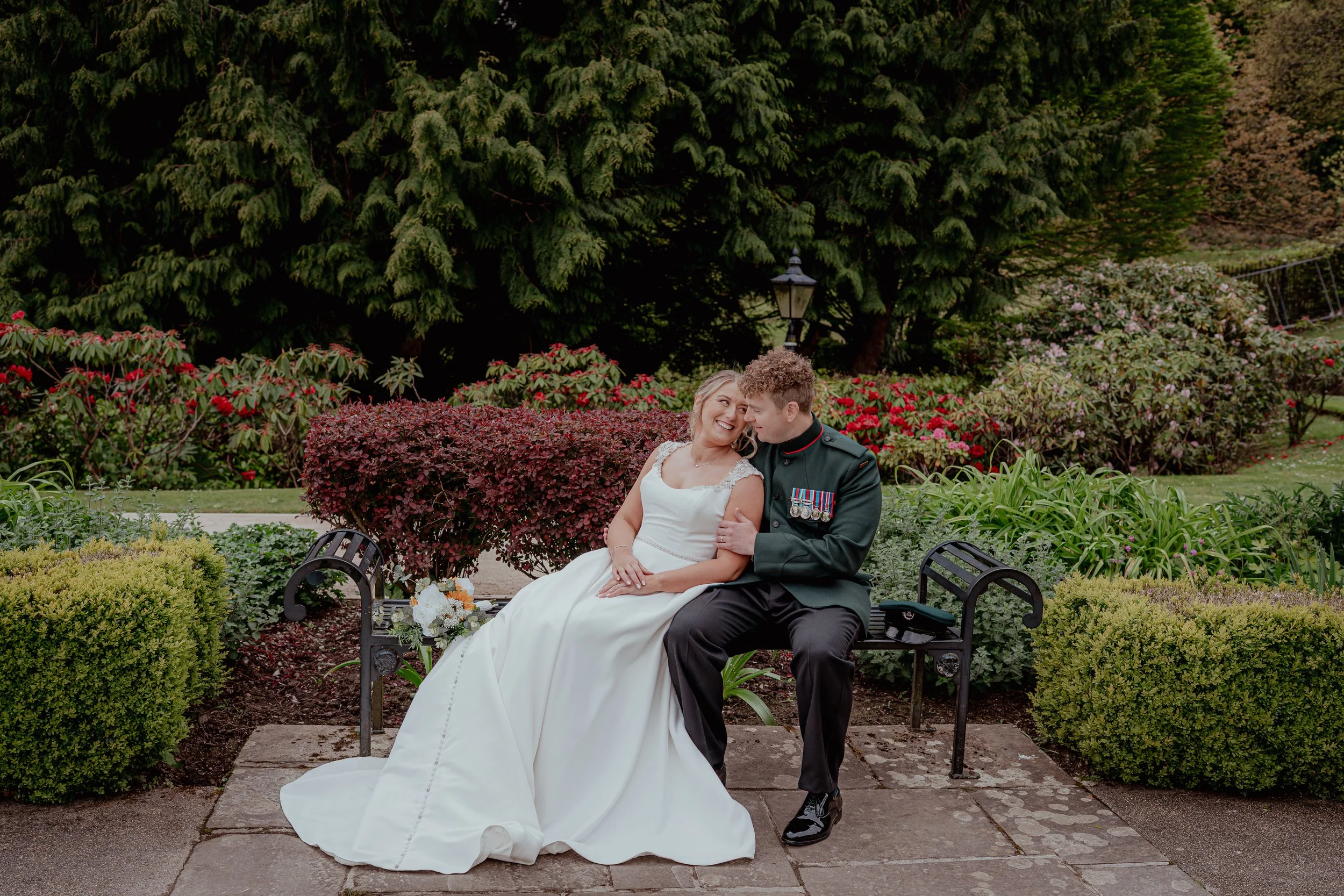 A bride and groom sitting on a park bench surrounded by lush greenery and colorful flowers, sharing a happy moment.