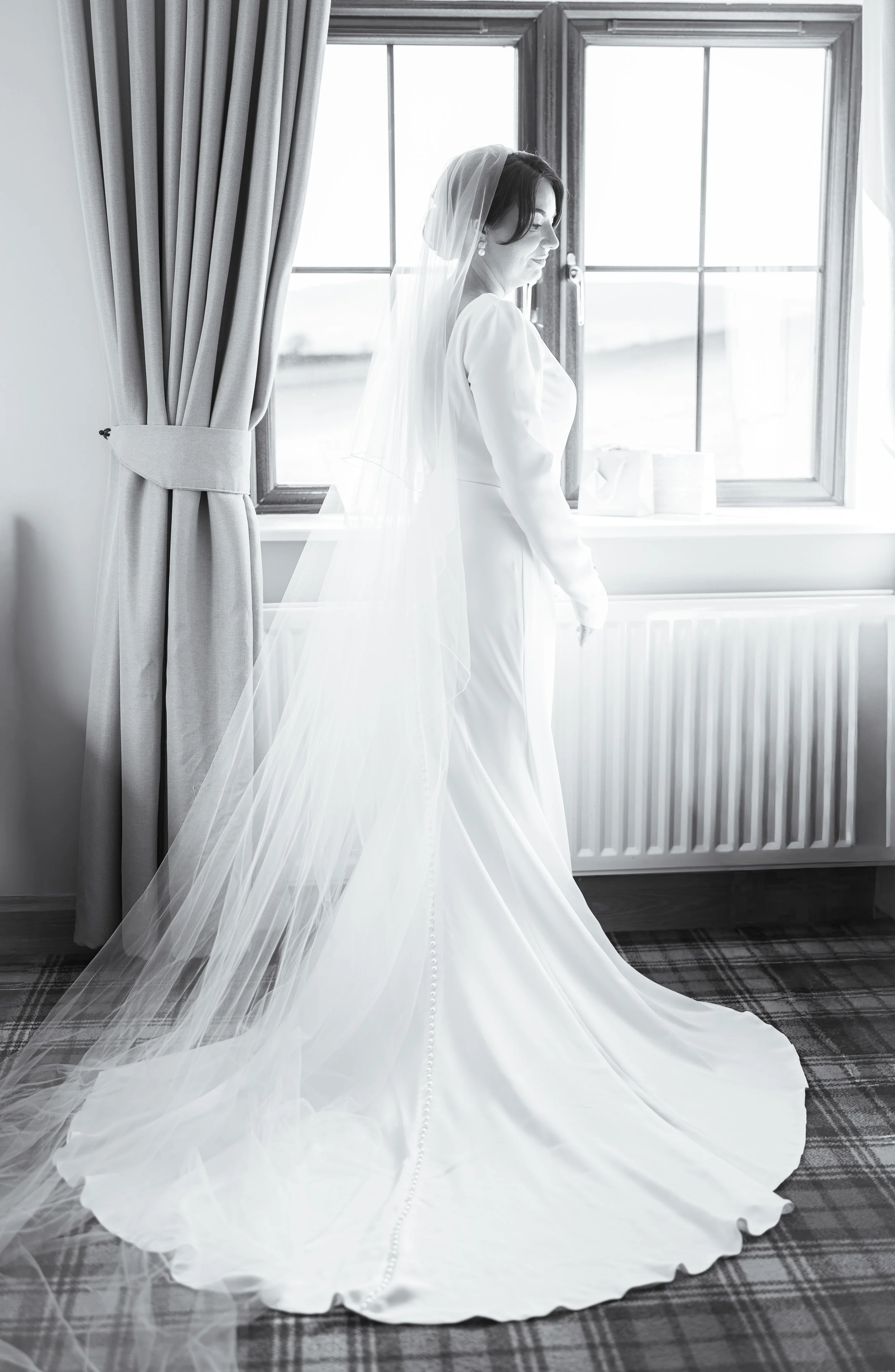 A bride in a white wedding dress and veil standing by a window, looking to the side.