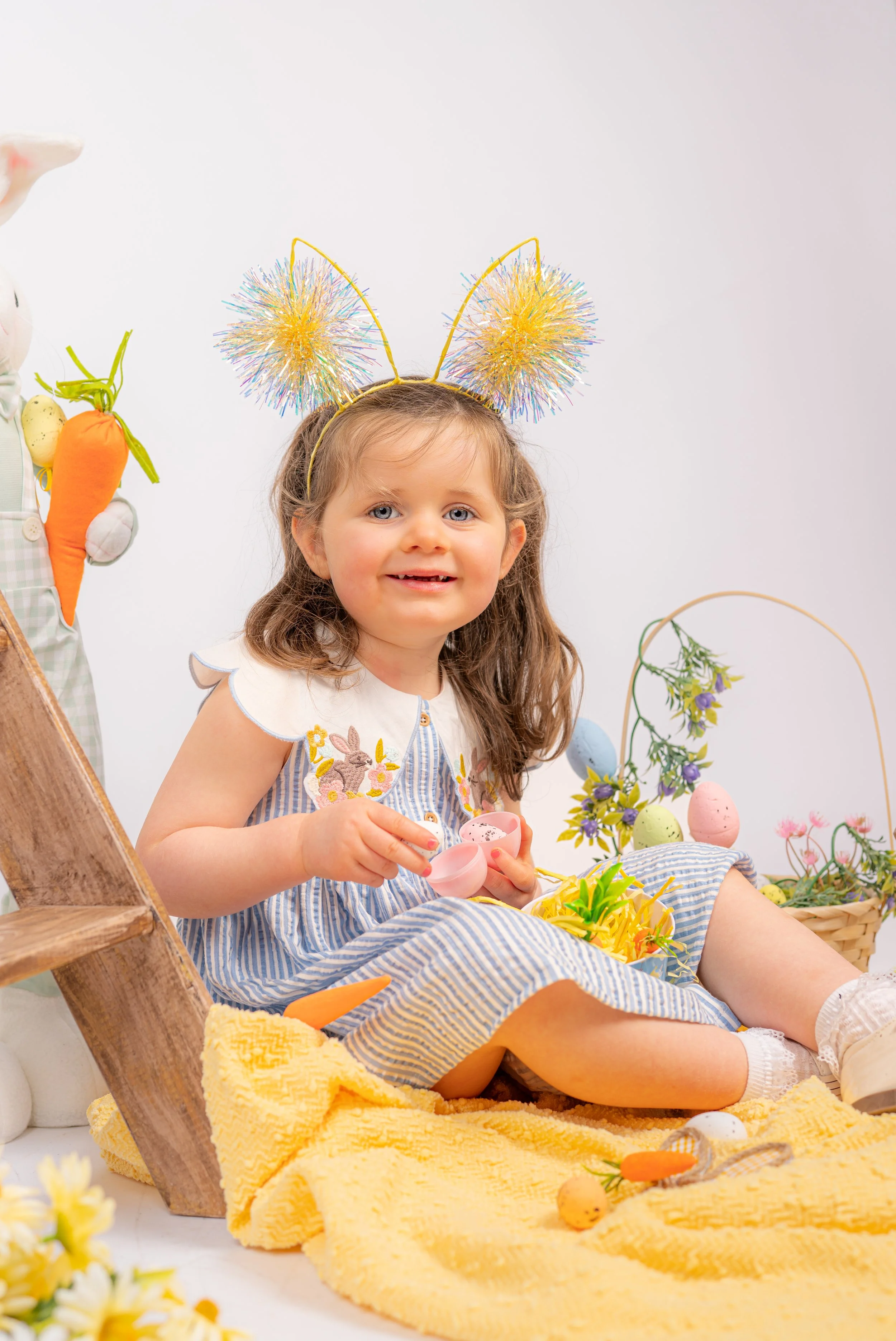 A young girl wearing bunny ears headband sits on a yellow blanket surrounded by Easter decorations, including pastel-colored eggs, a basket of flowers, and plush carrots, with a white background.