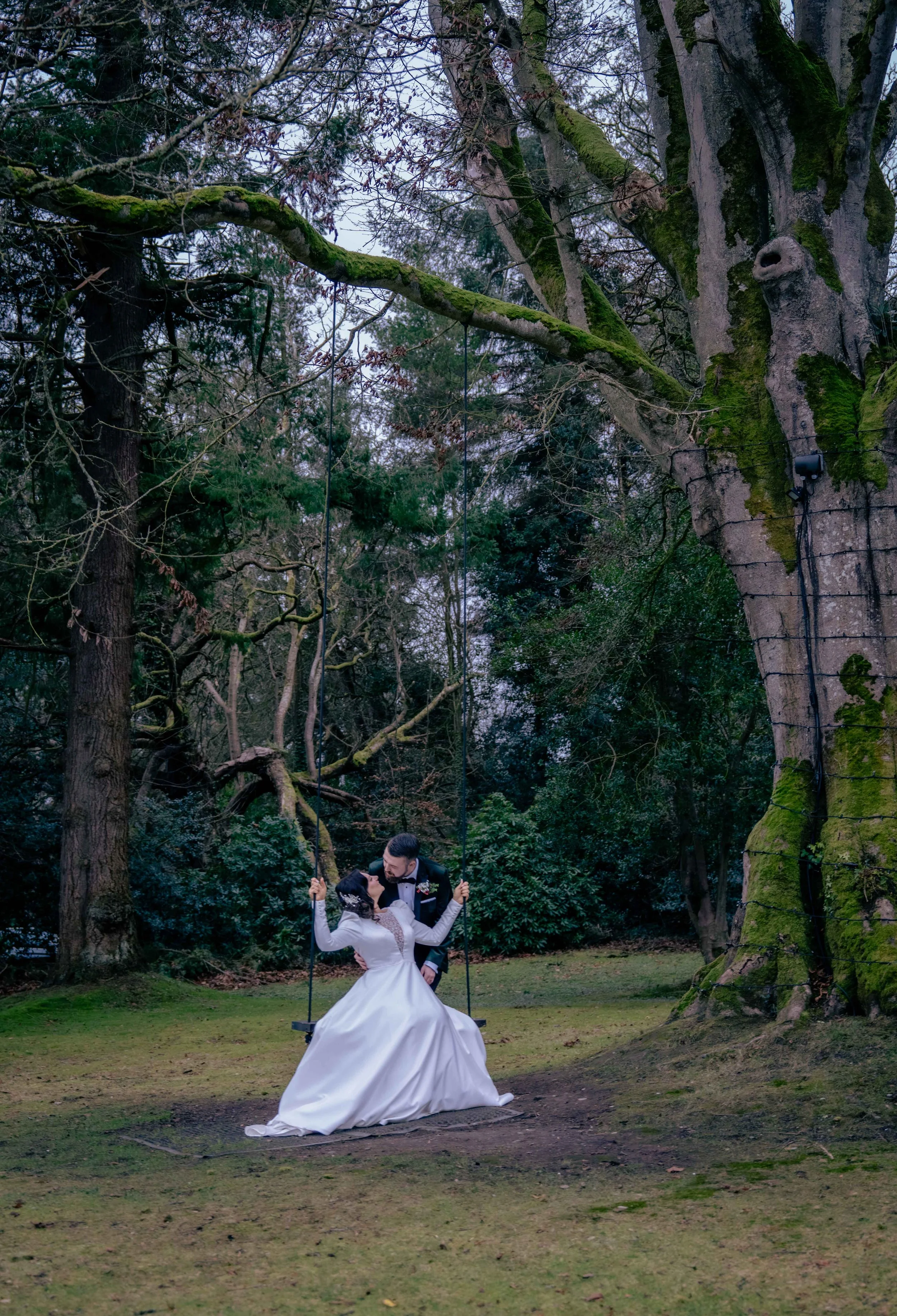 A bride and groom in wedding attire are sitting on a swing hanging from a large tree in a forested outdoor setting, sharing a romantic moment.