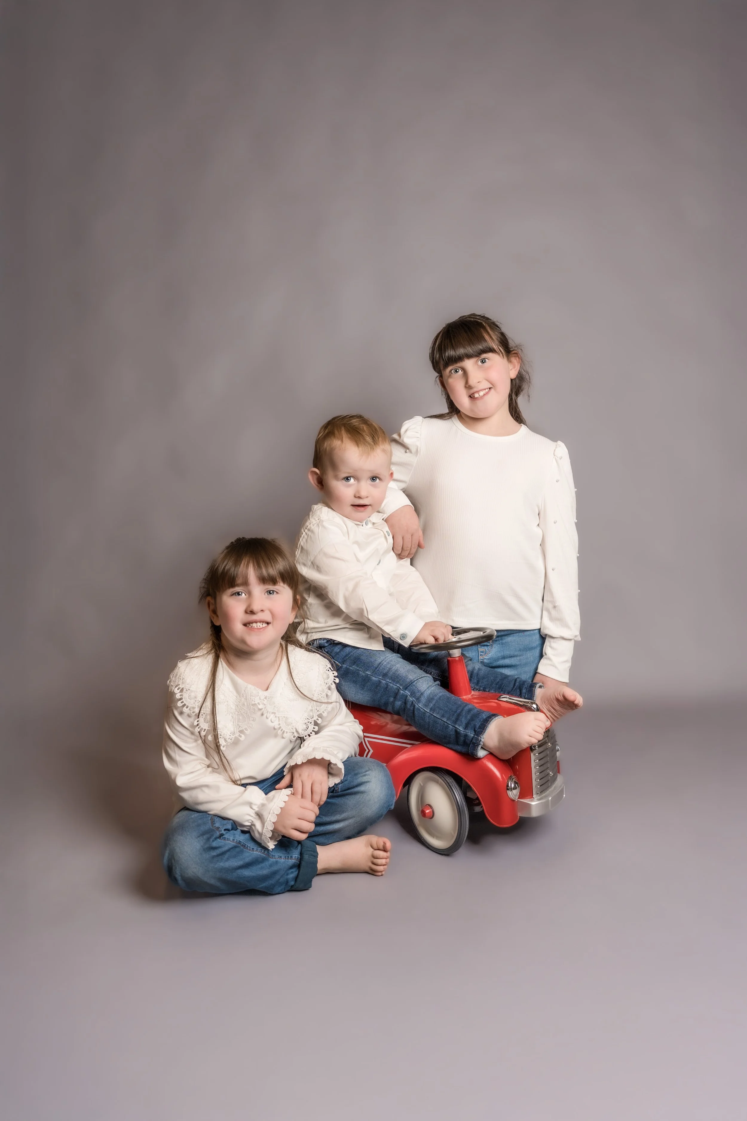 Three children, two girls and a boy, posing with a red toy fire truck in a studio setting with a gray backdrop.
