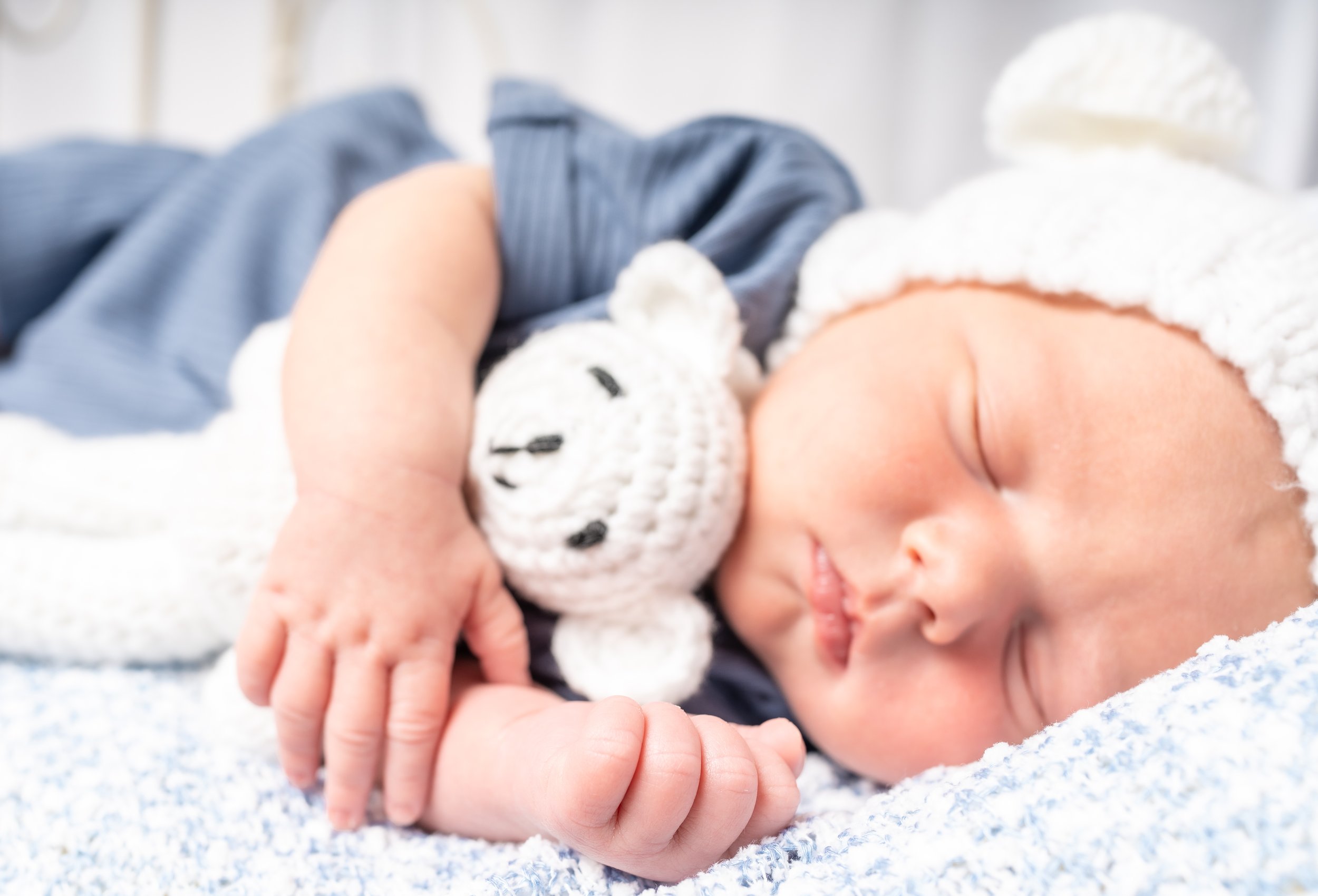 A sleeping baby lying on a blanket, wearing a knitted hat with a panda face and a blue outfit, holding a plush panda toy.