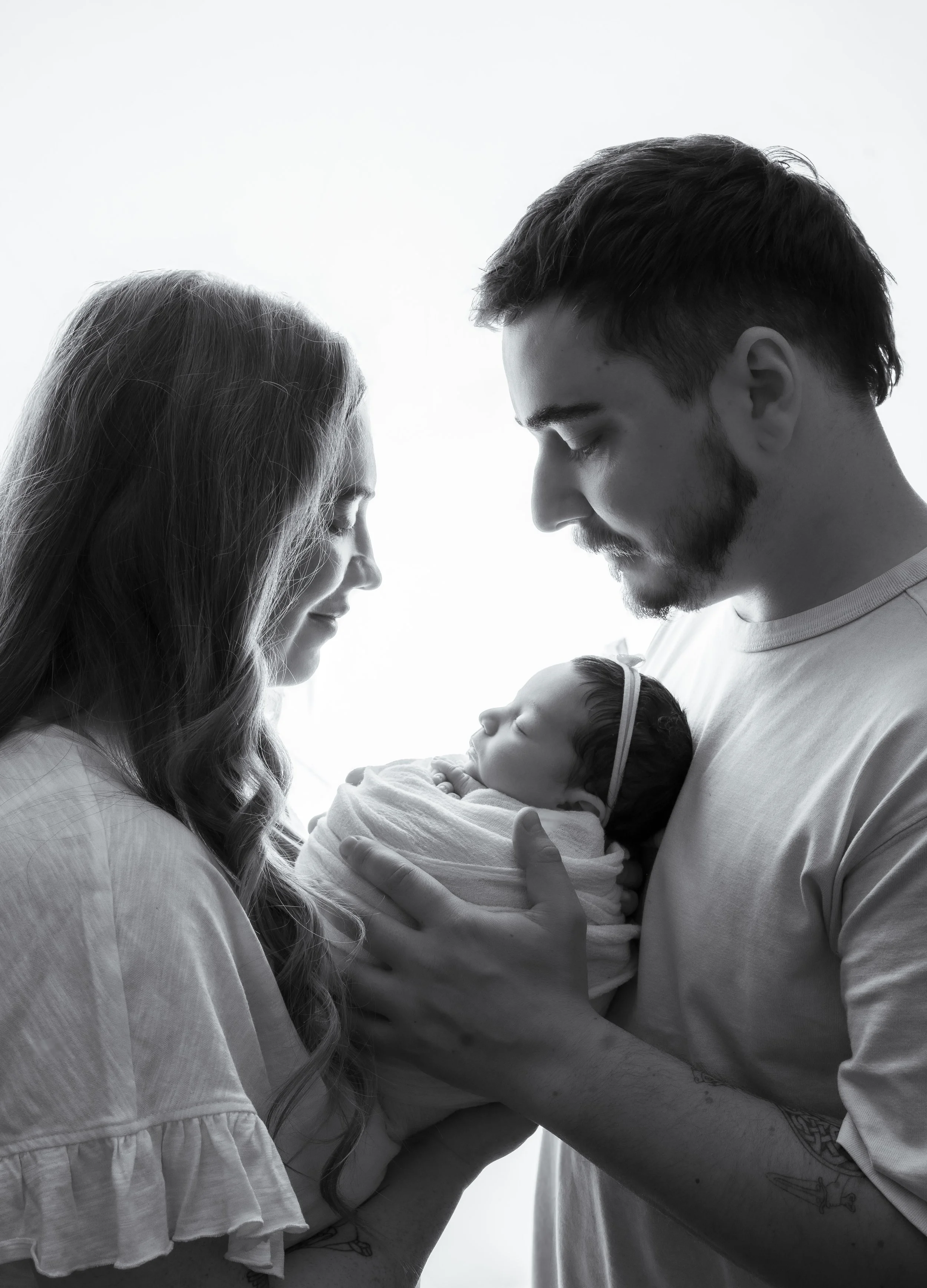 A black and white photograph of a family with a newborn. The woman and man are facing each other with their foreheads almost touching, both smiling softly, holding the baby between them. The woman has long wavy hair, and the man has short, dark hair and facial hair. The newborn is wrapped in a blanket and appears to be sleeping.