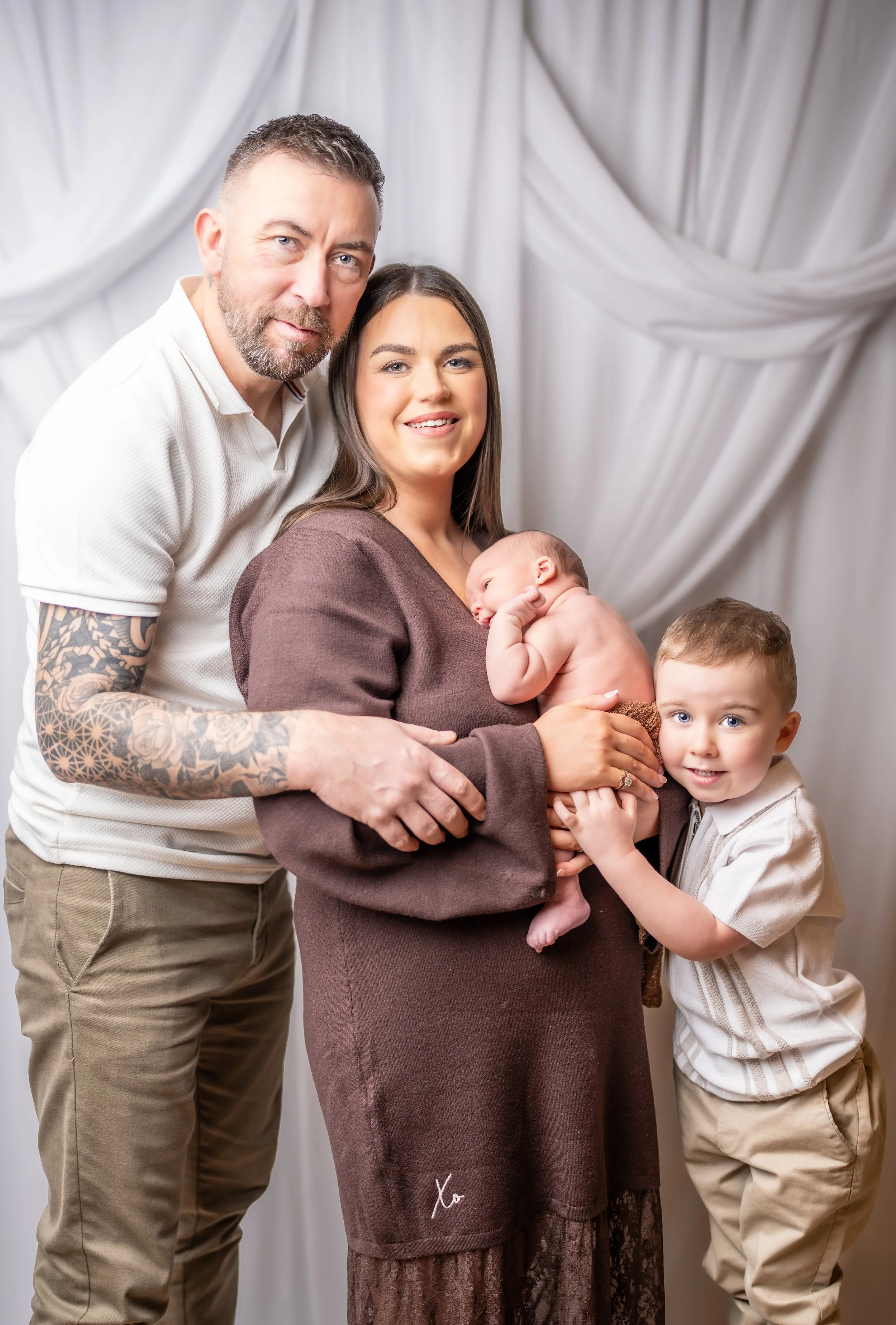 A family portrait of four family members, including a man with a beard and tattoos, a smiling woman holding a baby, and a young boy, standing in front of a white curtain backdrop.