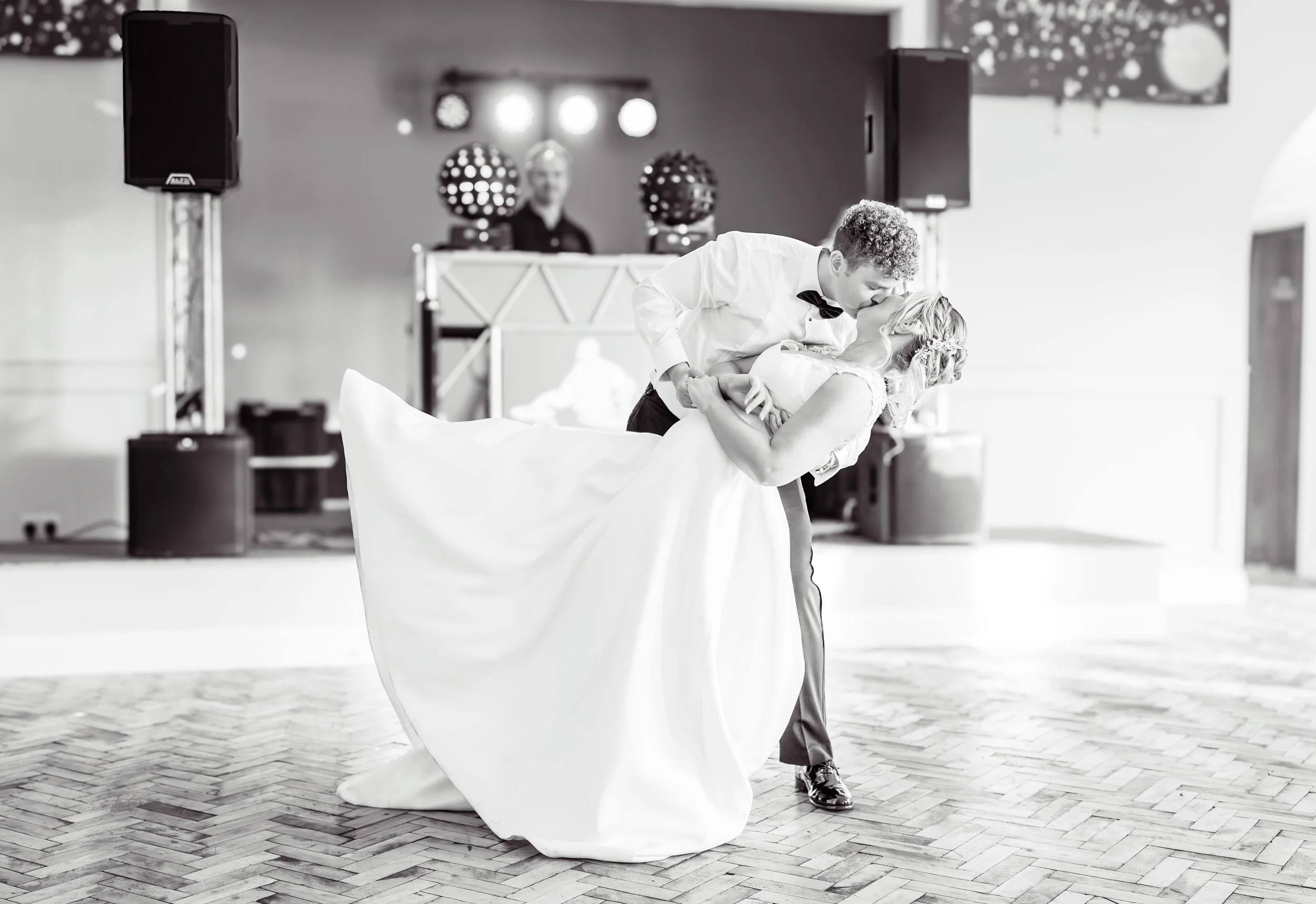 A bride and groom sharing a kiss during their wedding dance at a reception hall, with a DJ and sound equipment visible in the background.
