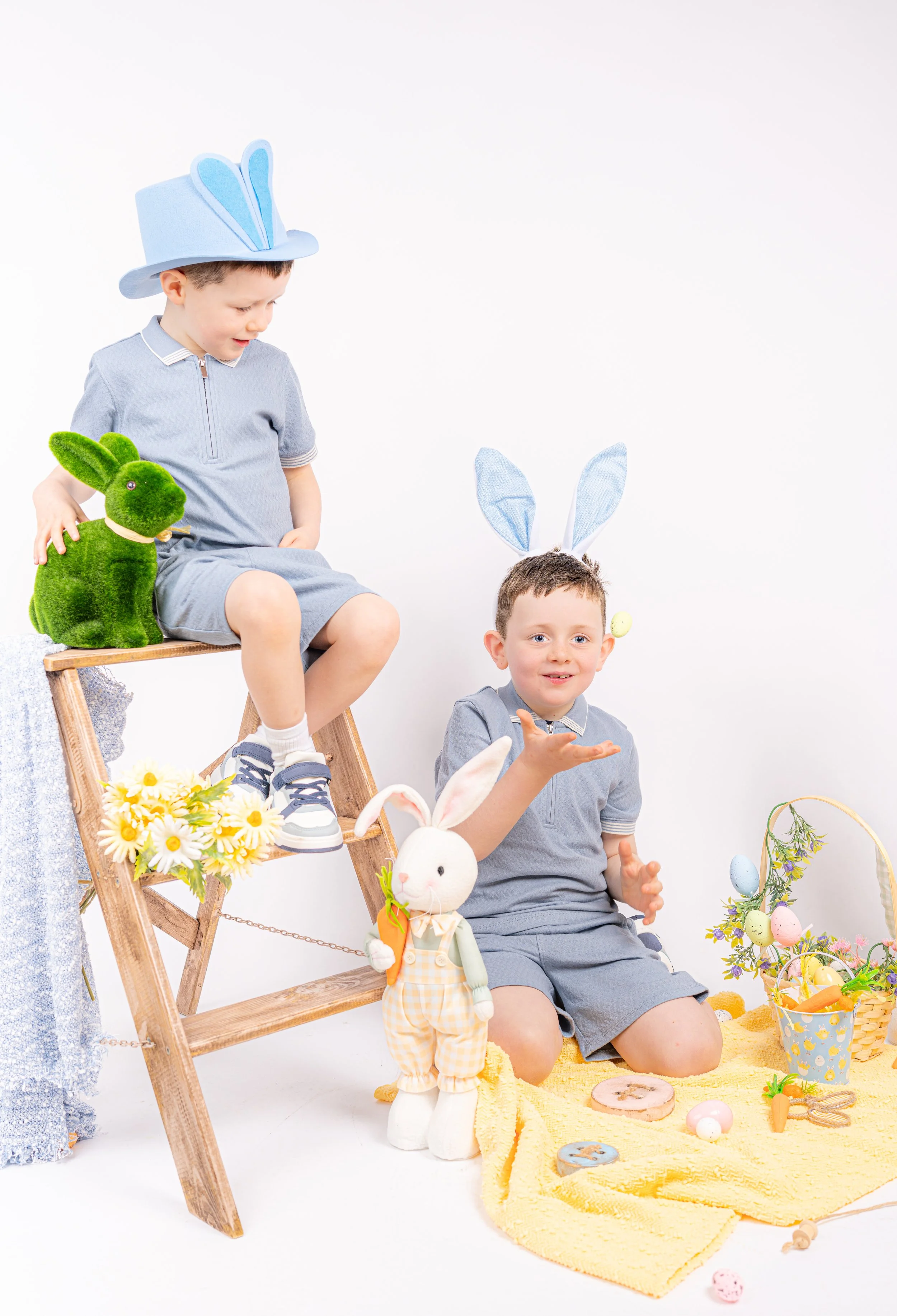 Two young boys dressed in gray with bunny ears, one sitting on a ladder and the other kneeling on the floor surrounded by Easter decorations and plush bunnies, celebrating Easter.