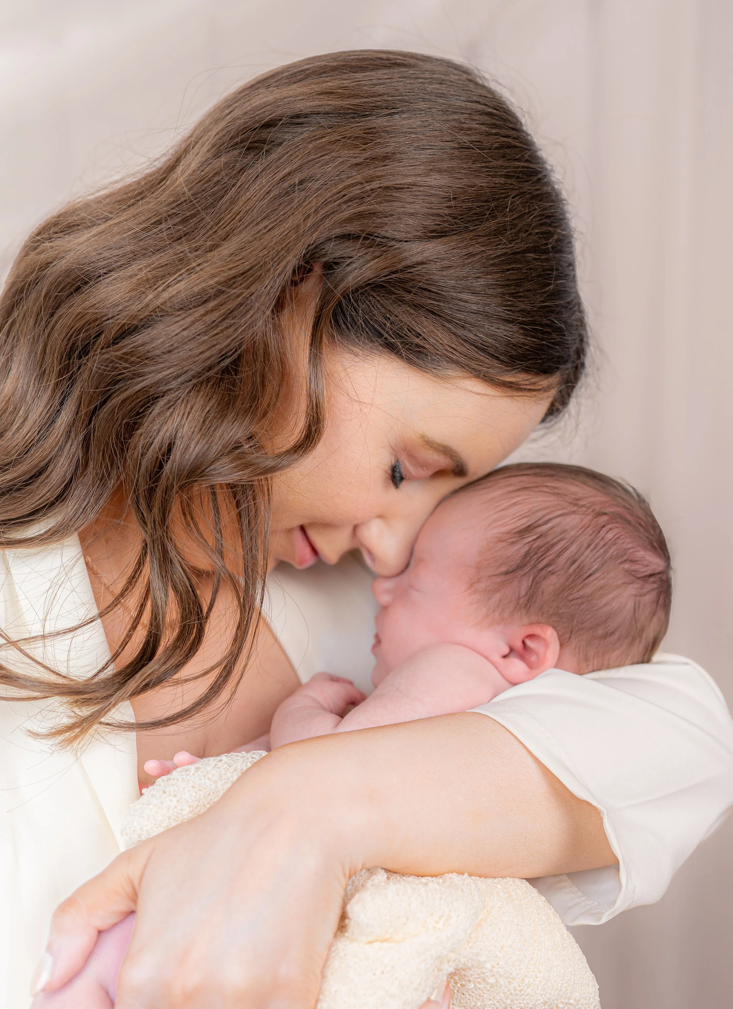 A woman with long, wavy brown hair is kissing a sleeping baby on the forehead. The woman is gently holding the baby close, and the baby is wrapped in a light-colored blanket.