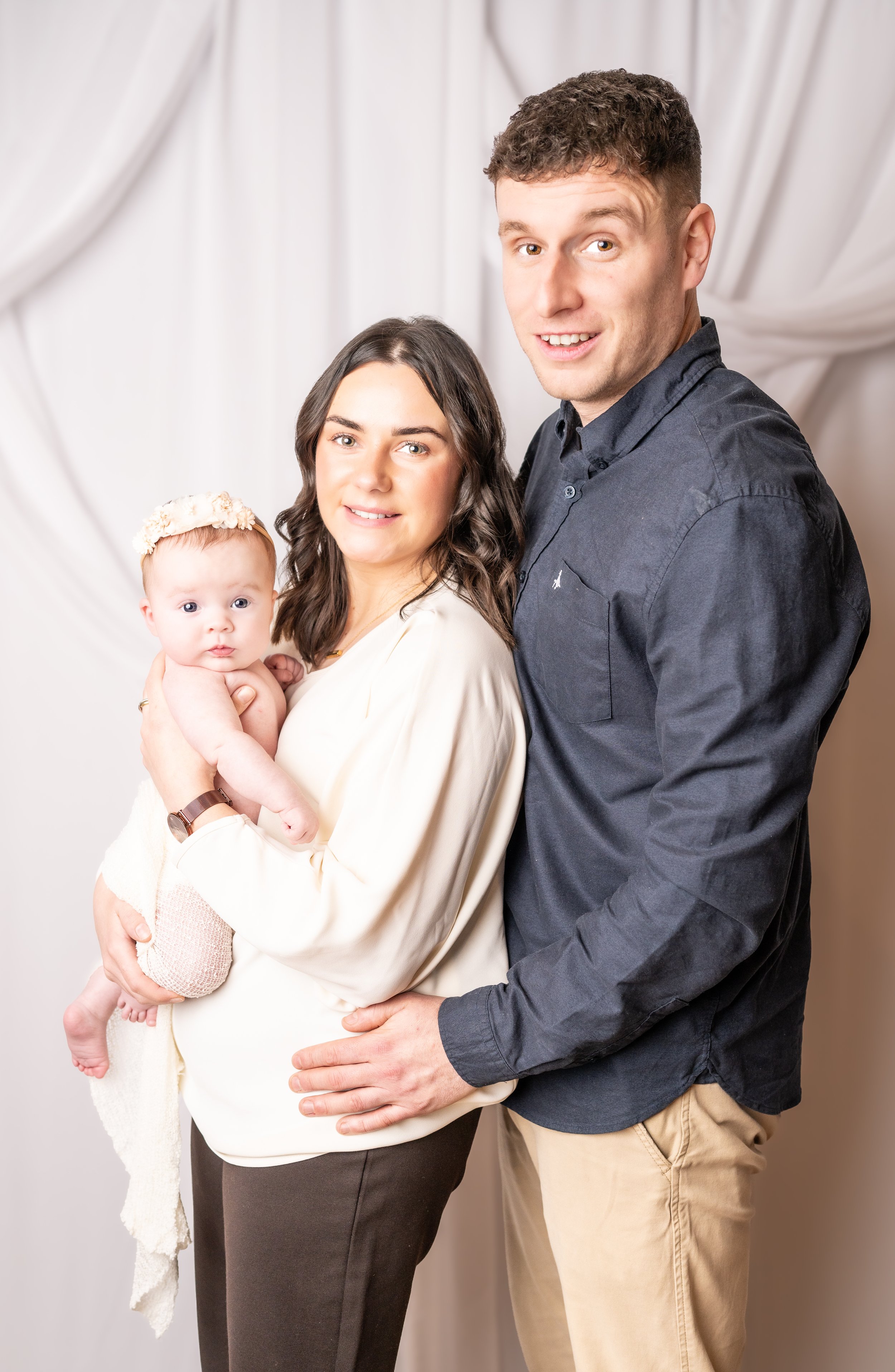 A woman holding a baby, standing between a man and a backdrop with white drapes.