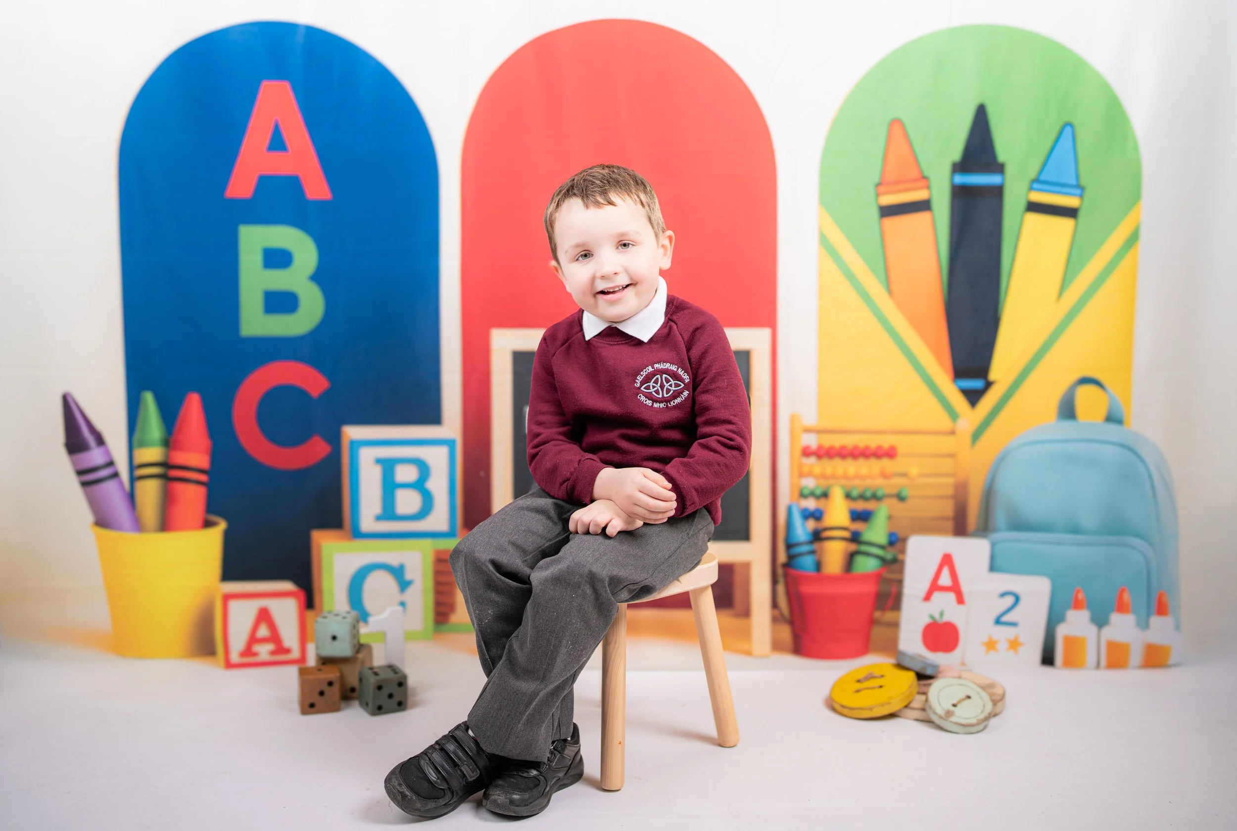 Young boy sitting on a small stool in front of colorful classroom decorations, including large alphabet letters, toy blocks, crayons, and educational materials, with a light-colored background.