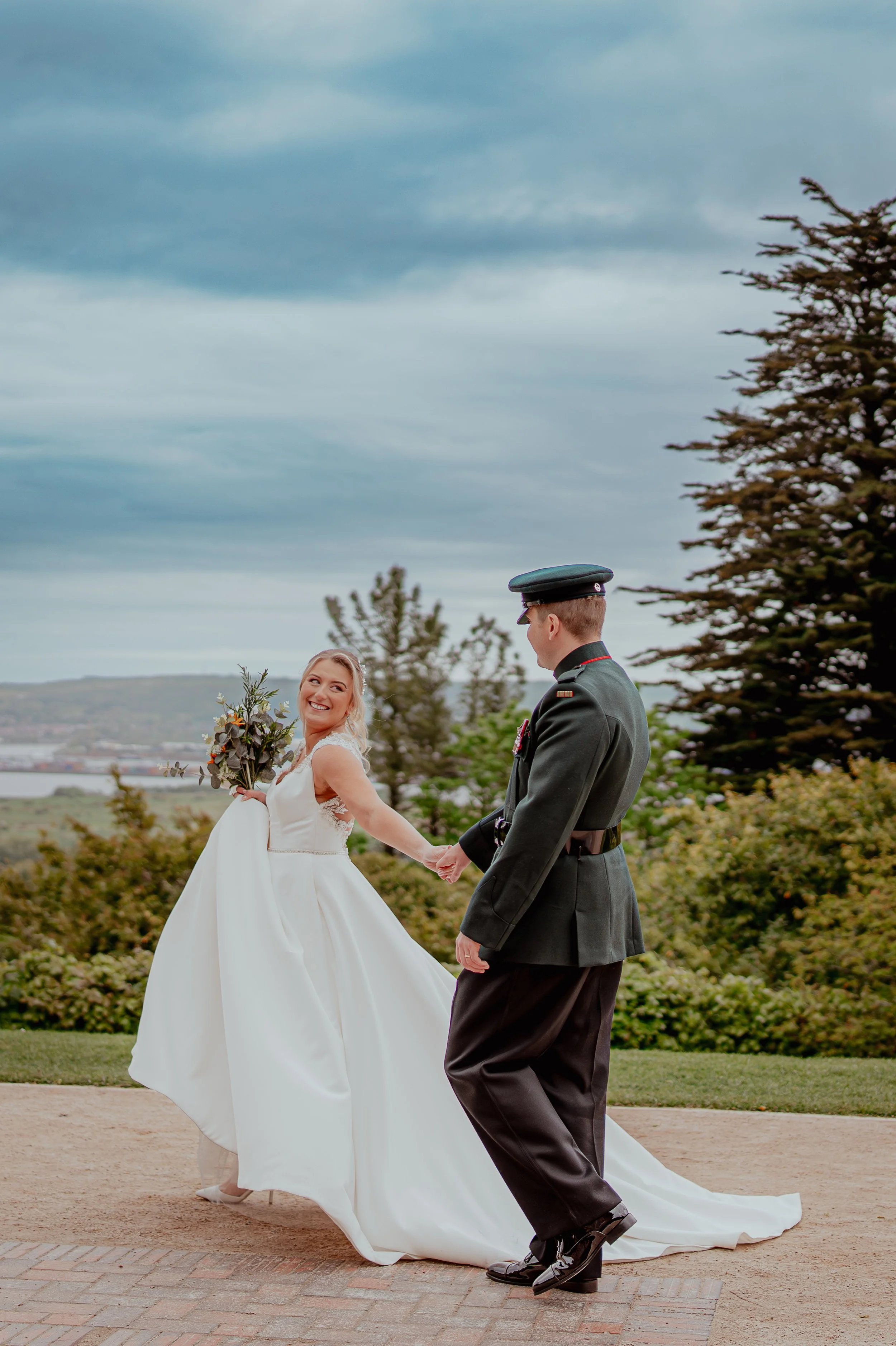 A bride and groom holding hands outdoors during a wedding ceremony. The bride is smiling and wearing a white wedding dress, holding a bouquet of flowers. The groom is dressed in a dark military uniform and a military cap. The background features trees, cloudy sky, and a hilly landscape.
