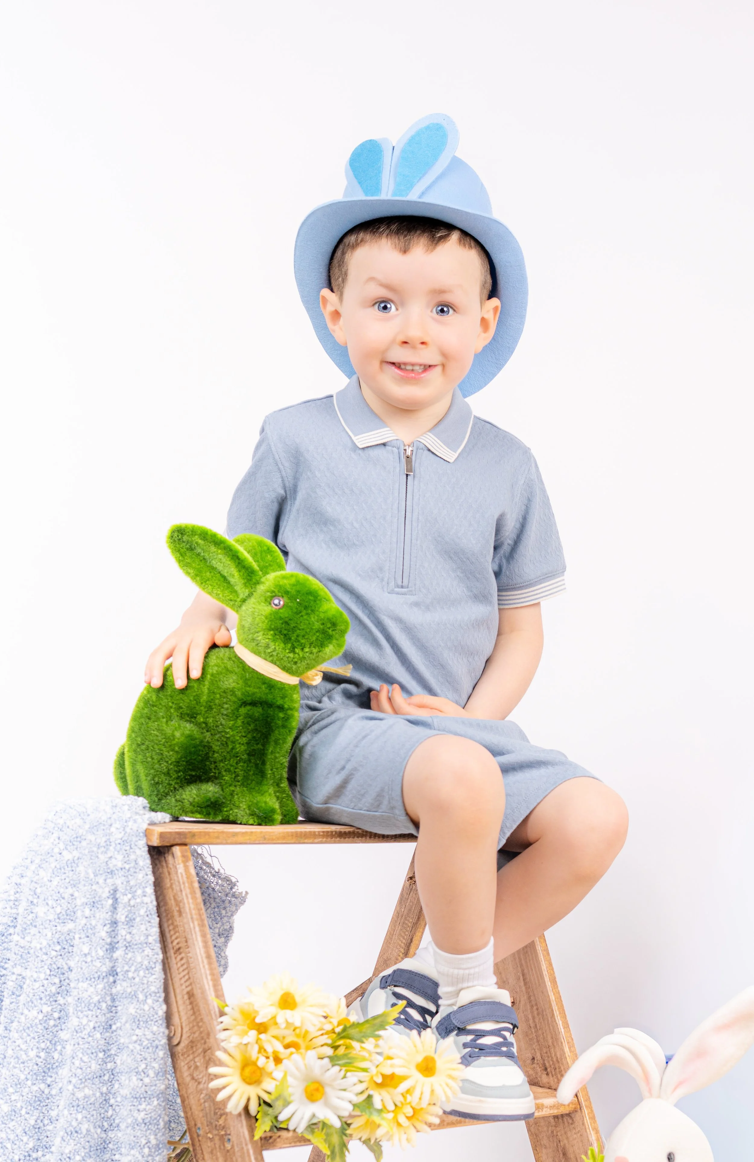 A young boy dressed for Easter, wearing a blue hat with bunny ears, a light blue zip-up shirt, and shorts, is sitting on a wooden ladder. He has a green plush bunny on his lap and is smiling. There are yellow daisies in front of him and a white stuffed bunny partially visible to the right.