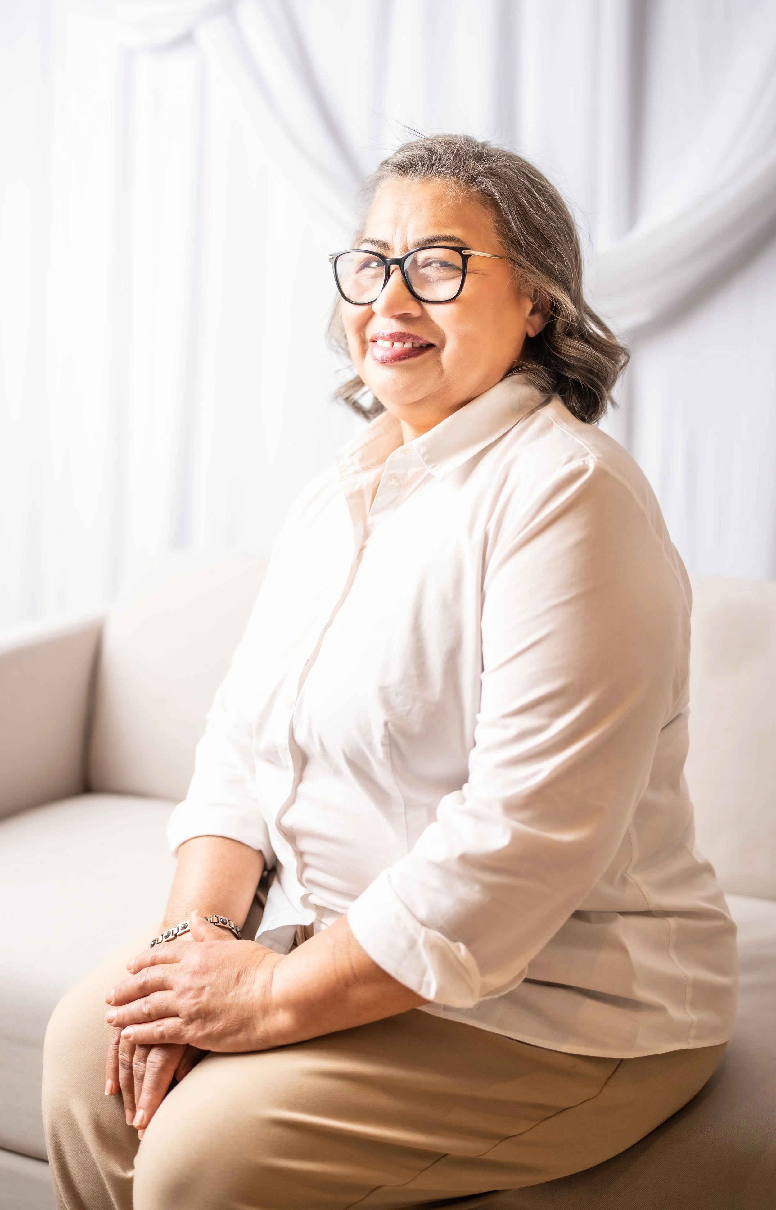 A middle-aged woman with gray hair and glasses sitting on a beige couch, smiling, with white curtains in the background.
