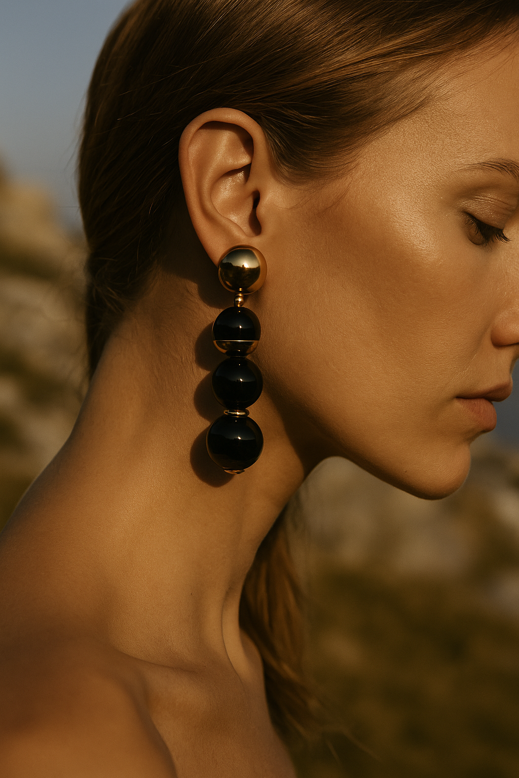 Close-up de uma mulher com pele clara, cabelo castanho claro, usando brincos grandes dourados e pretos, ao ar livre ao entardecer, com fundo desfocado de uma paisagem natural.