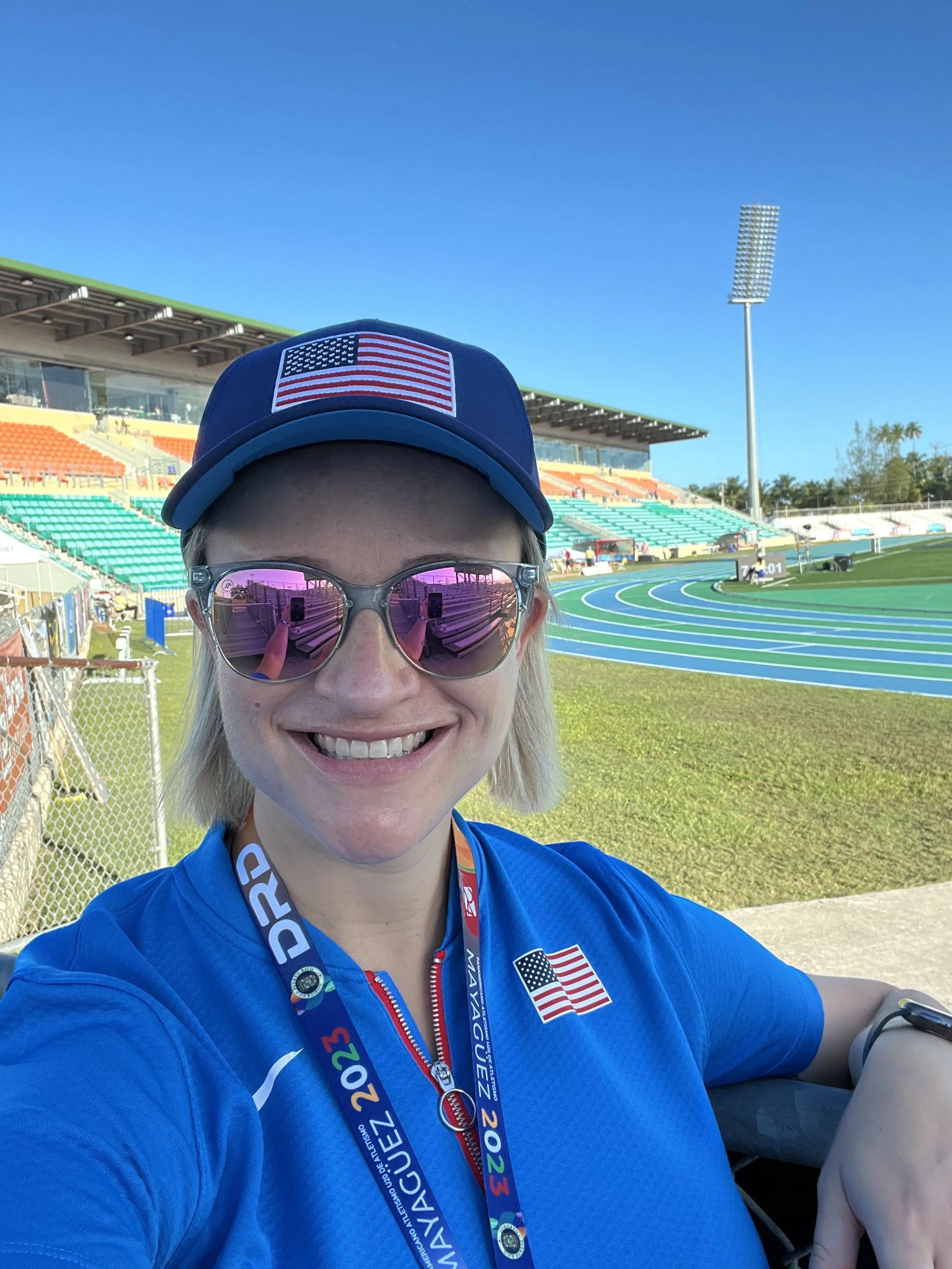 Dr. Erin Wos, a smiling woman in sunglasses and a blue shirt wearing a medal and a cap with the American flag at a sports stadium, with track lanes and empty seats in the background.
