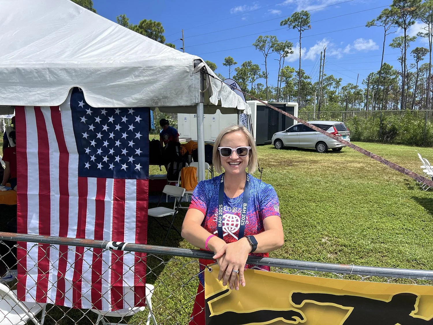 Dr. Erin Wos, a woman with sunglasses standing outdoors at an event, smiling, with a tent, American flag, and cars in the background.