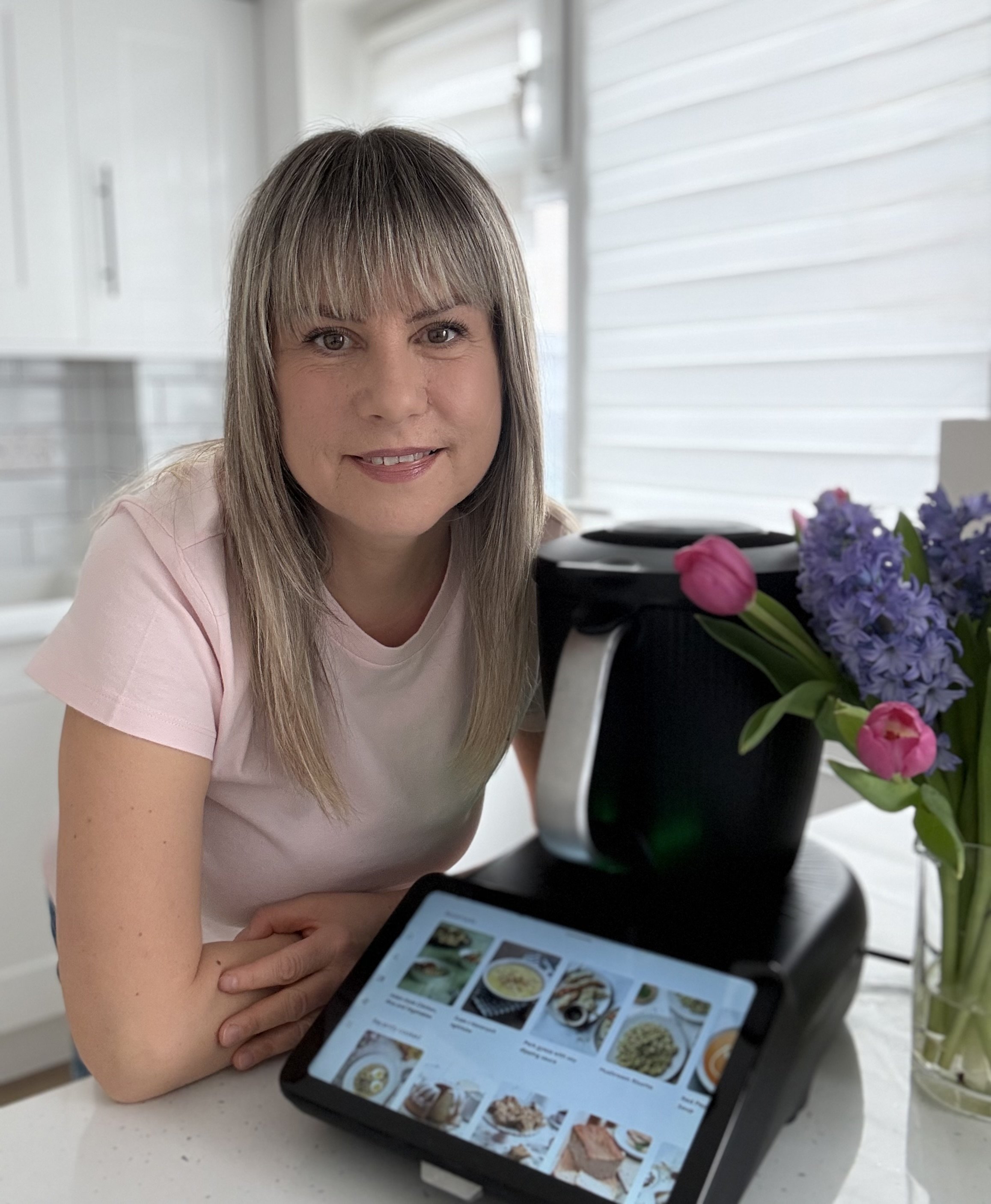 A woman with long blonde hair and bangs, wearing a light pink shirt, sitting at a table with a tablet displaying a menu of food items, holding her phone and looking at the camera with a slight smile. There are flowers in a black pot on the table, and the background shows a window with white blinds.