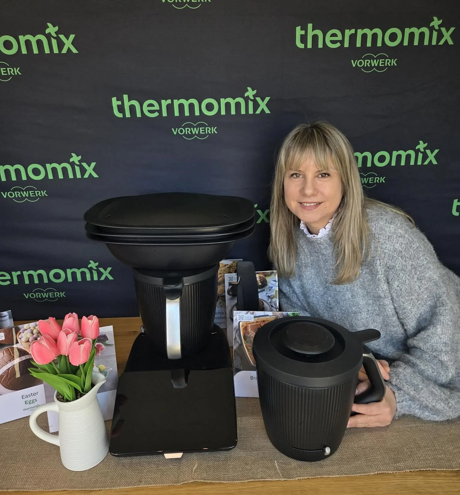 A woman is smiling and sitting at a table with kitchen appliance Thermomix TM7 , including a black air fryer and a small black Thermomix TM7  in front of a backdrop with green text that reads "thermomix" and "VORWERK."