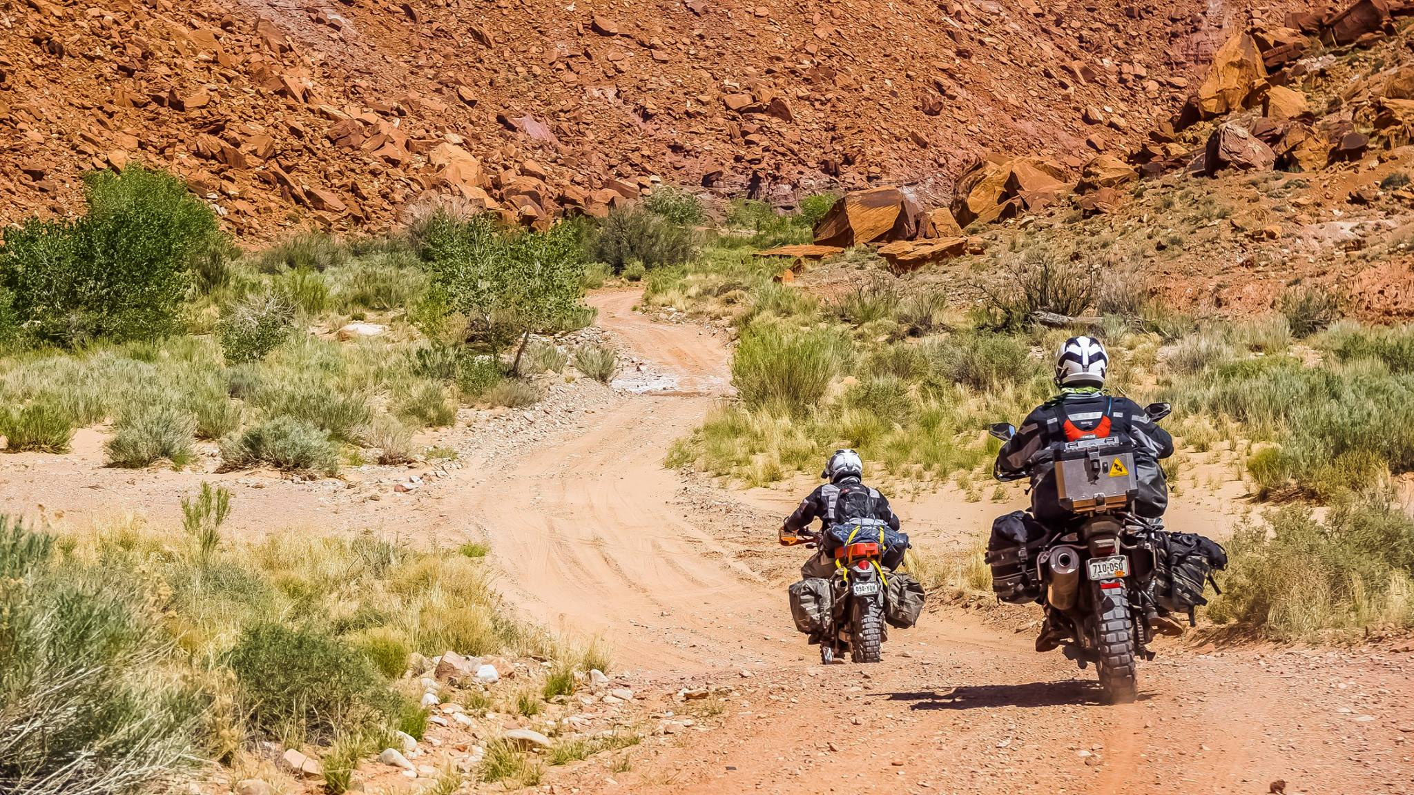 Two motorcyclists riding on a dirt trail through a desert landscape with sparse vegetation and red rocky hills in the background.