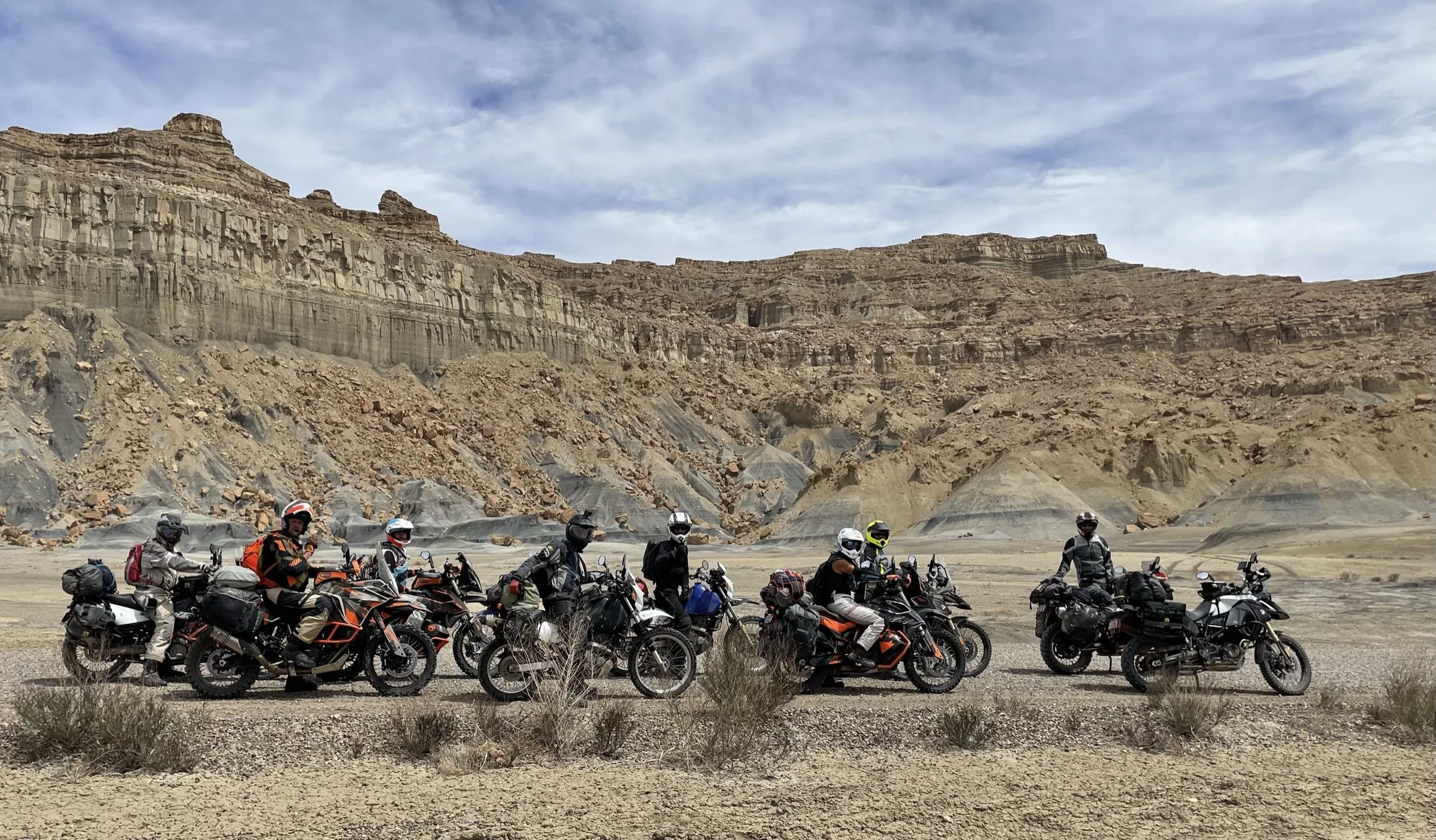 A group of motorcyclists with gear and helmets riding off-road bikes in a desert landscape with rocky cliffs and sparse vegetation.
