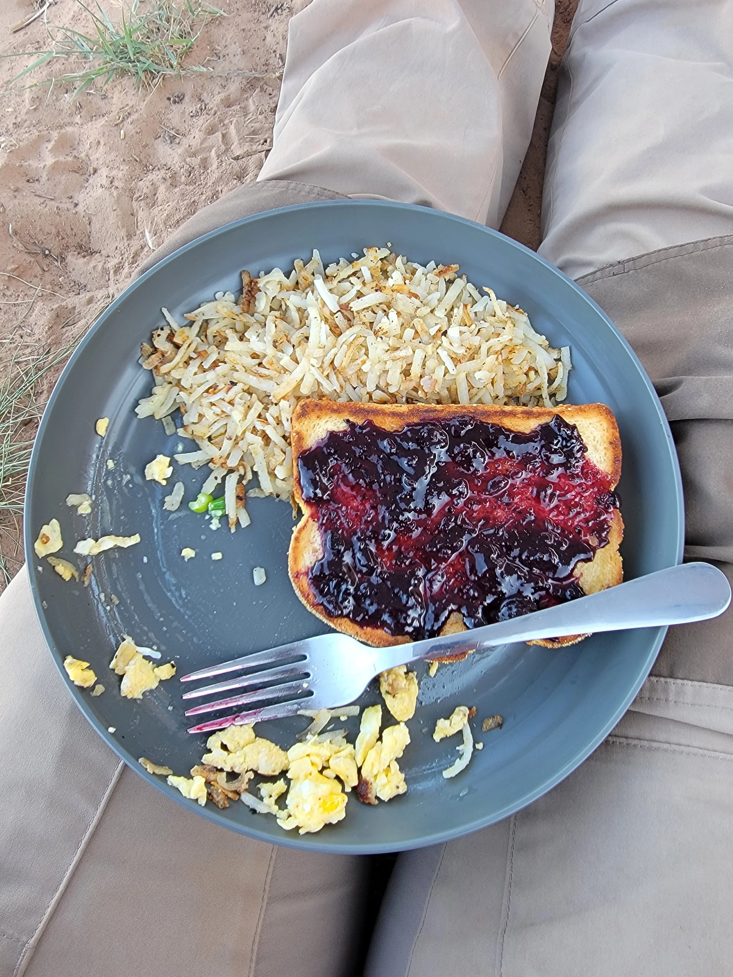 A gray plate on a person's lap contains scrambled eggs, hash browns, and a slice of toast with mixed berry jam.