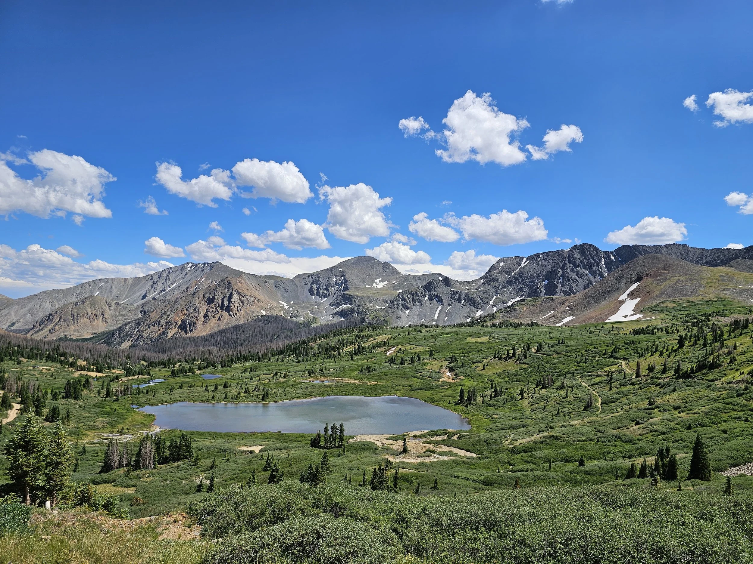 Scenic mountain landscape with a lake, green vegetation, and snow patches on rugged mountain peaks under a partly cloudy blue sky.