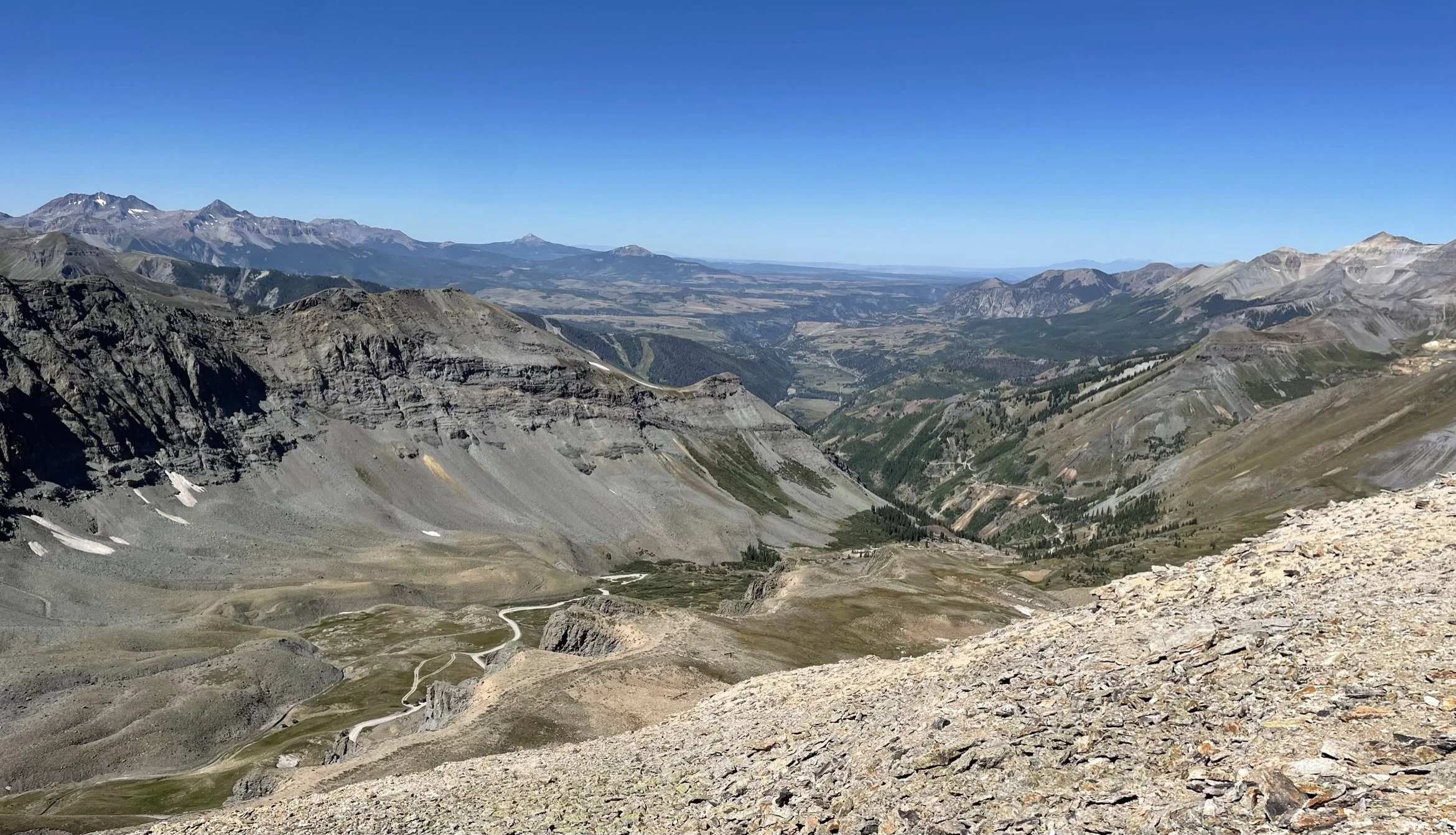 A vast mountain landscape with rugged slopes, winding roads, green patches, and a blue sky.