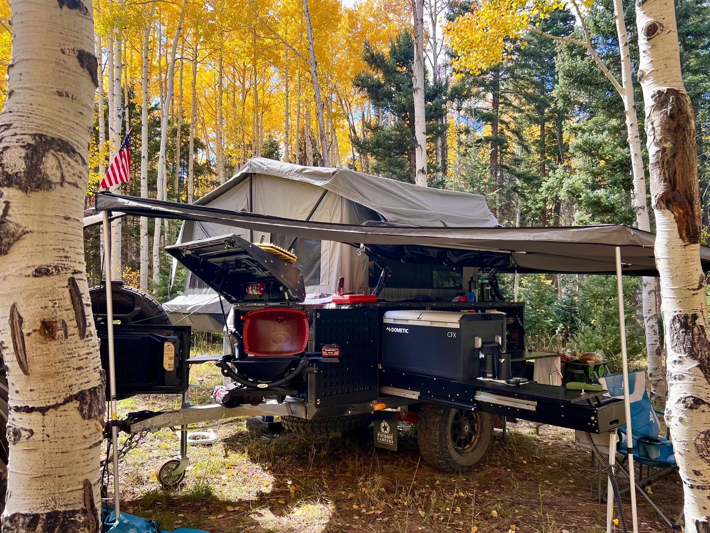 Camping setup in a forest with trees, a trailer with an awning, and outdoor cooking gear.