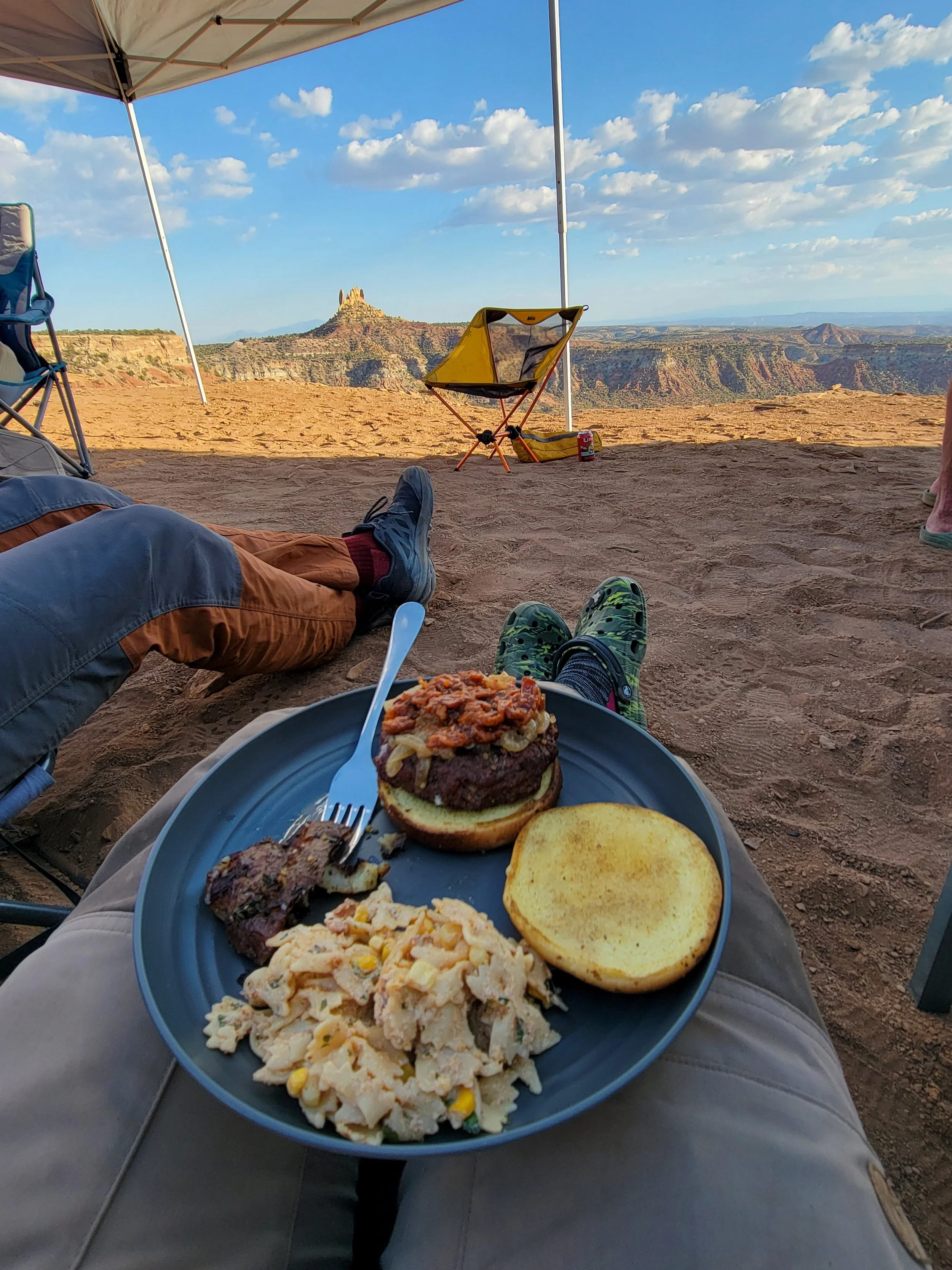 Person sitting outdoors under a canopy, enjoying a meal with a view of canyons and a mountain in the background, with a plate of food containing a burger, macaroni salad, a potato pancake, and grilled meat, in a scenic desert landscape.